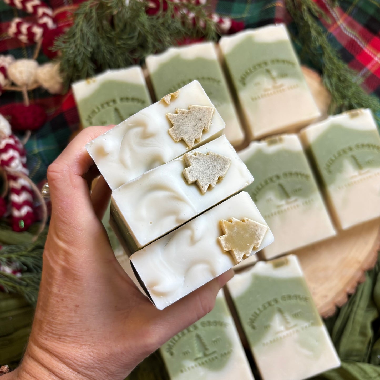 Hand holding a block of soap with Christmas tree designs, surrounded by more soap bars on a festive background.