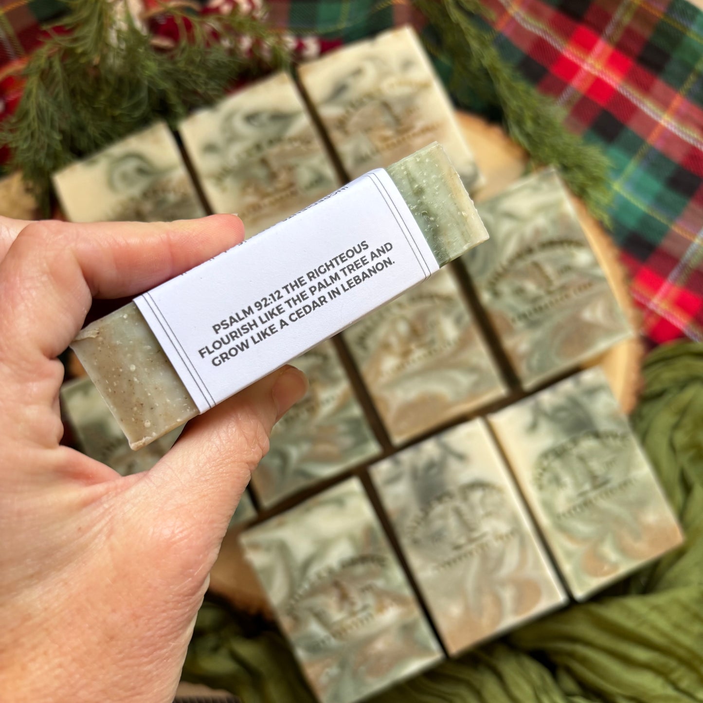 Hand holding a soap bar with a label in front of more soap bars on a plaid fabric background.