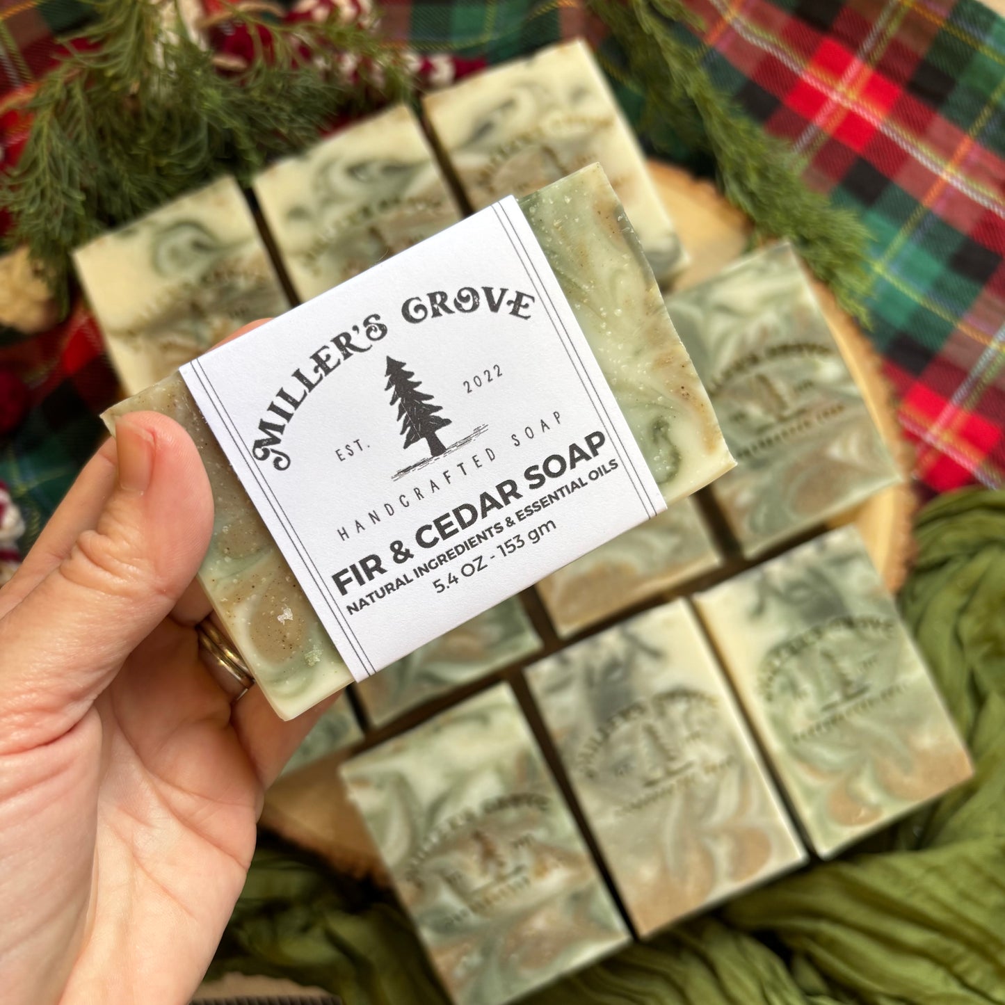 Handcrafted soap bar with 'Miller's Grove' label held by a hand, surrounded by more soap bars and greenery.