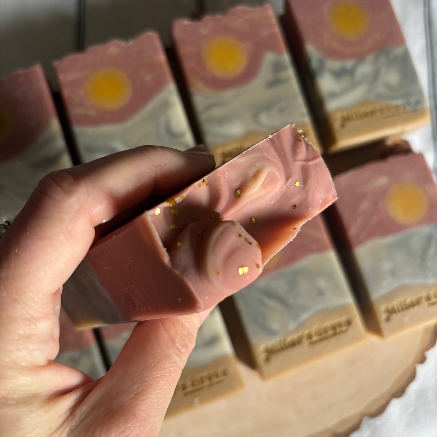 Hand holding a pink soap bar with gold specks, with more soap bars in the background.