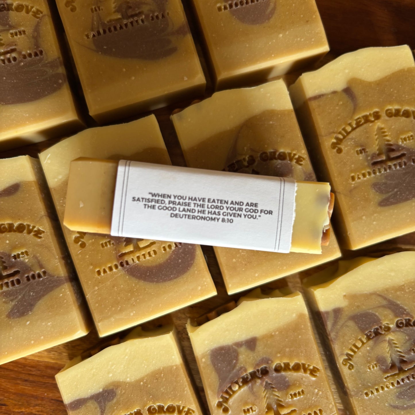 A close-up of handmade rectangular bars of soap arranged on a wooden surface, with a white paper label wrapped around one bar. The soap has a warm cream and tan marbled look with darker brown swirls, and the embossed text reads '"Miller's Grove"