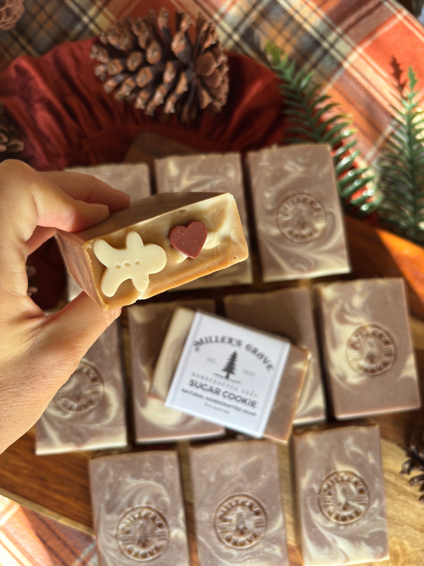 A hand holds a decorated bar of soap in the shape of a sugar cookie, with several other marbled soap bars arranged on a wooden surface in the background. A label reads 'Miller's Grove Sugar Cookie'.