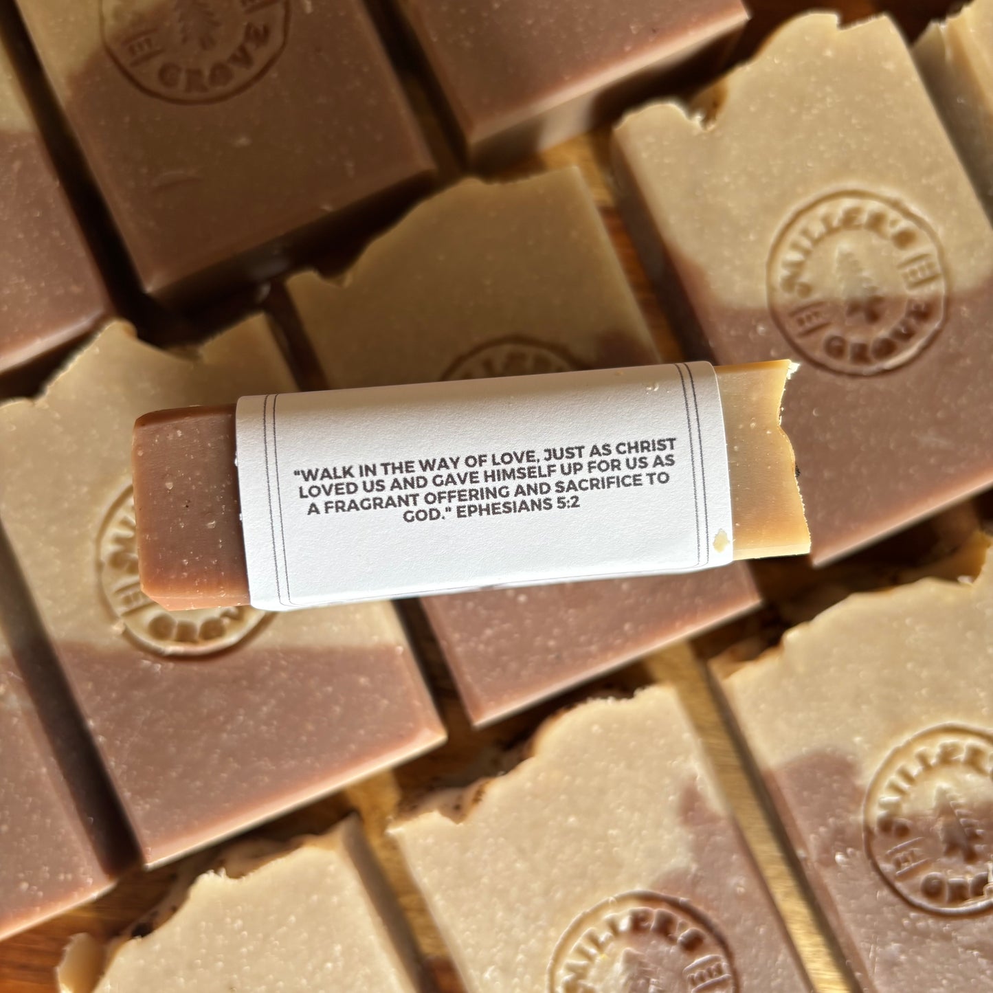 A close-up of a rectangular stick of butter soap lying horizontally on top of several stacked butter soaps. The central soap has a white paper label wrapped around it with printed black text, and the surrounding soaps are light tan and medium brown with embossed circular logos.