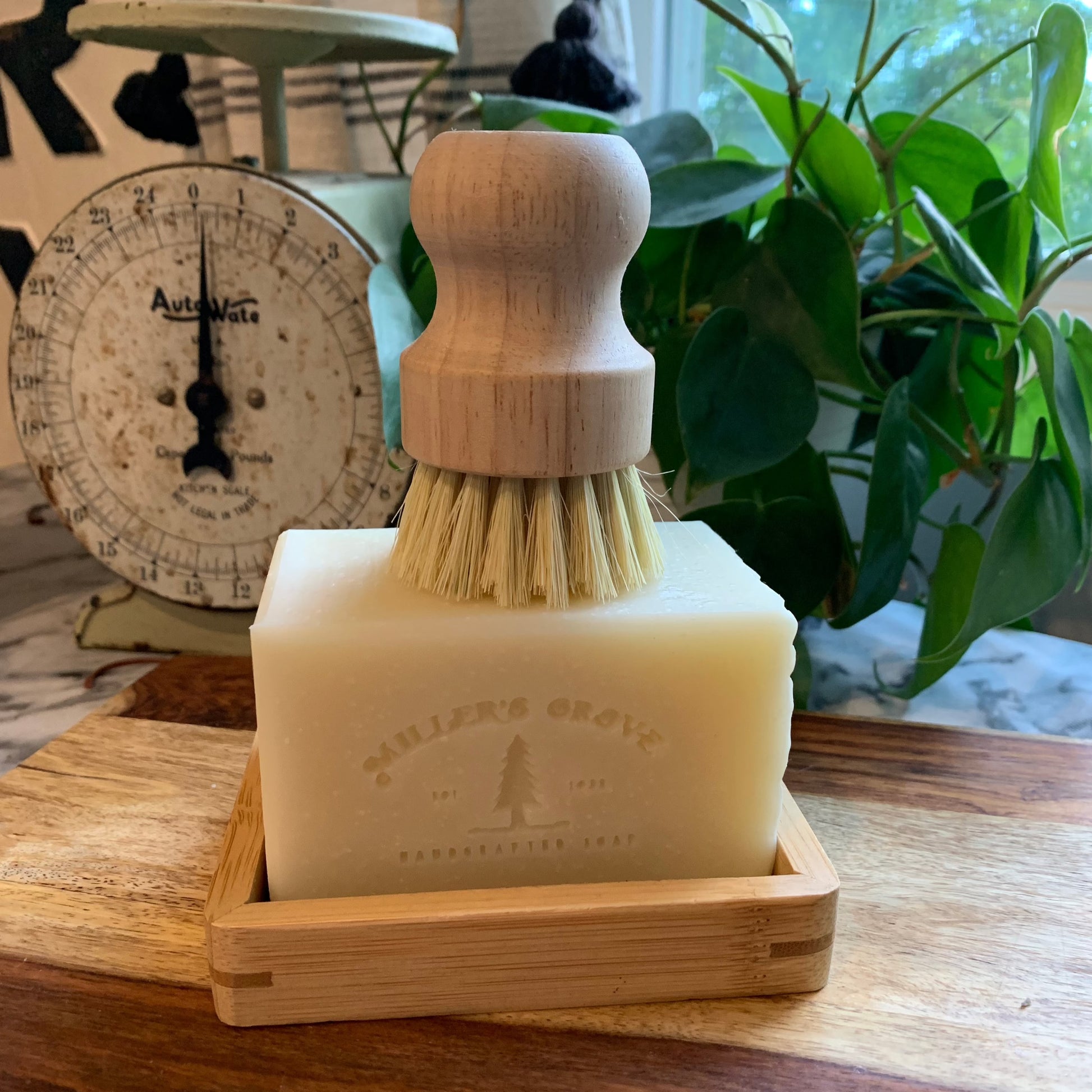 beige rectangular brick of soap on a wooden soap tray with a wood handled scrubber brush on top