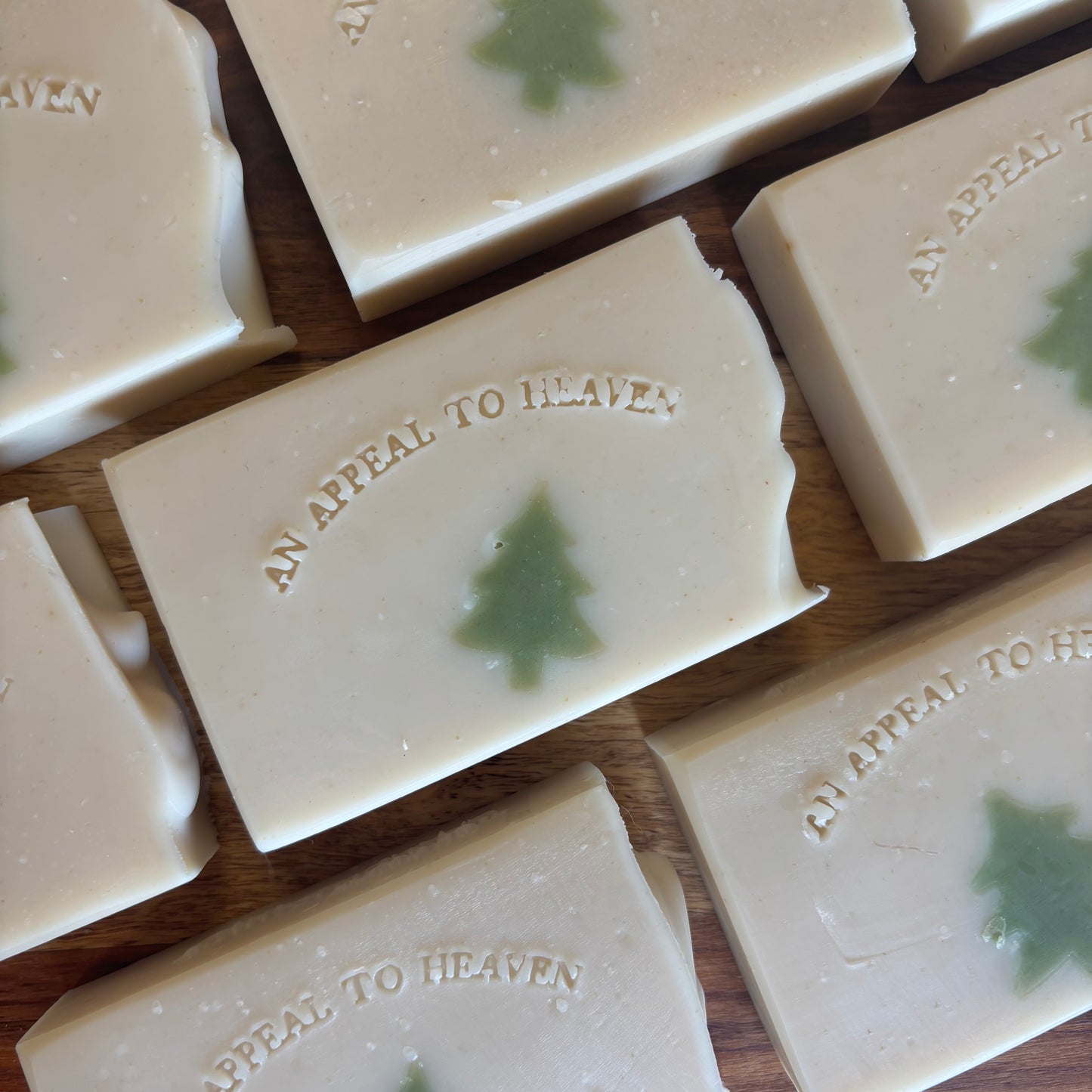 Several rectangular bars of soap arranged on a wooden surface. The soap is a warm off-white/cream color with a small green Christmas tree motif in the center and the words 'AN APPEAL TO HEAVEN' embossed on the top.