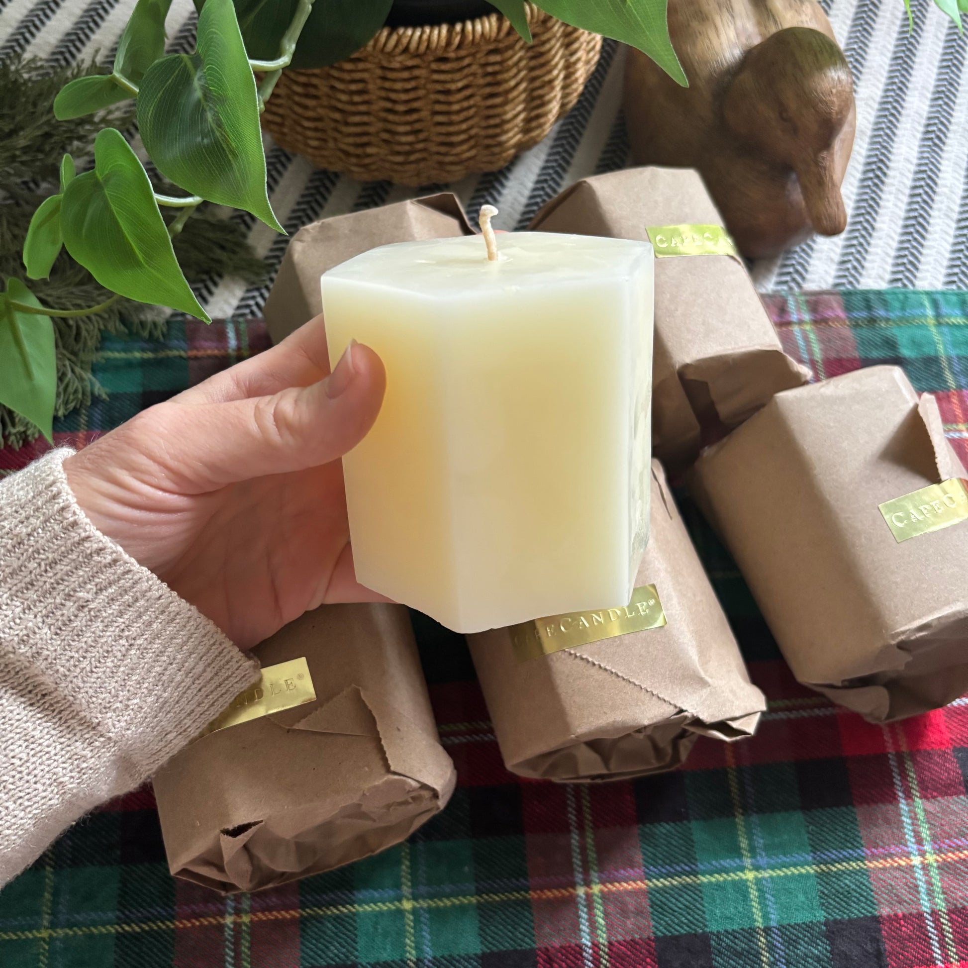Hand holding a wrapped candle with more candles in brown paper packaging on a plaid surface.