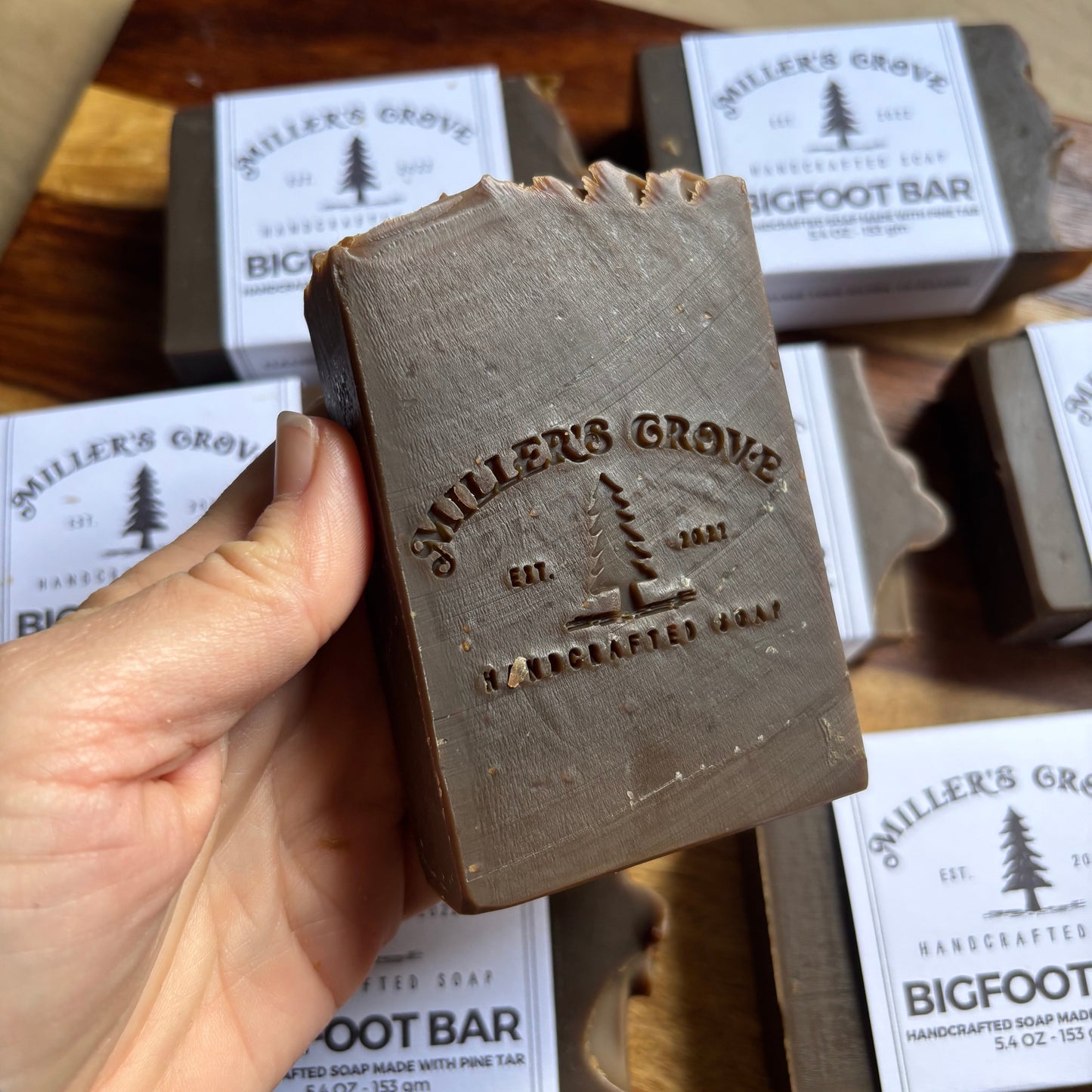 Hand holding a bar of soap with 'Wilder's Grove' branding, surrounded by more soap bars and packaging on a wooden surface.