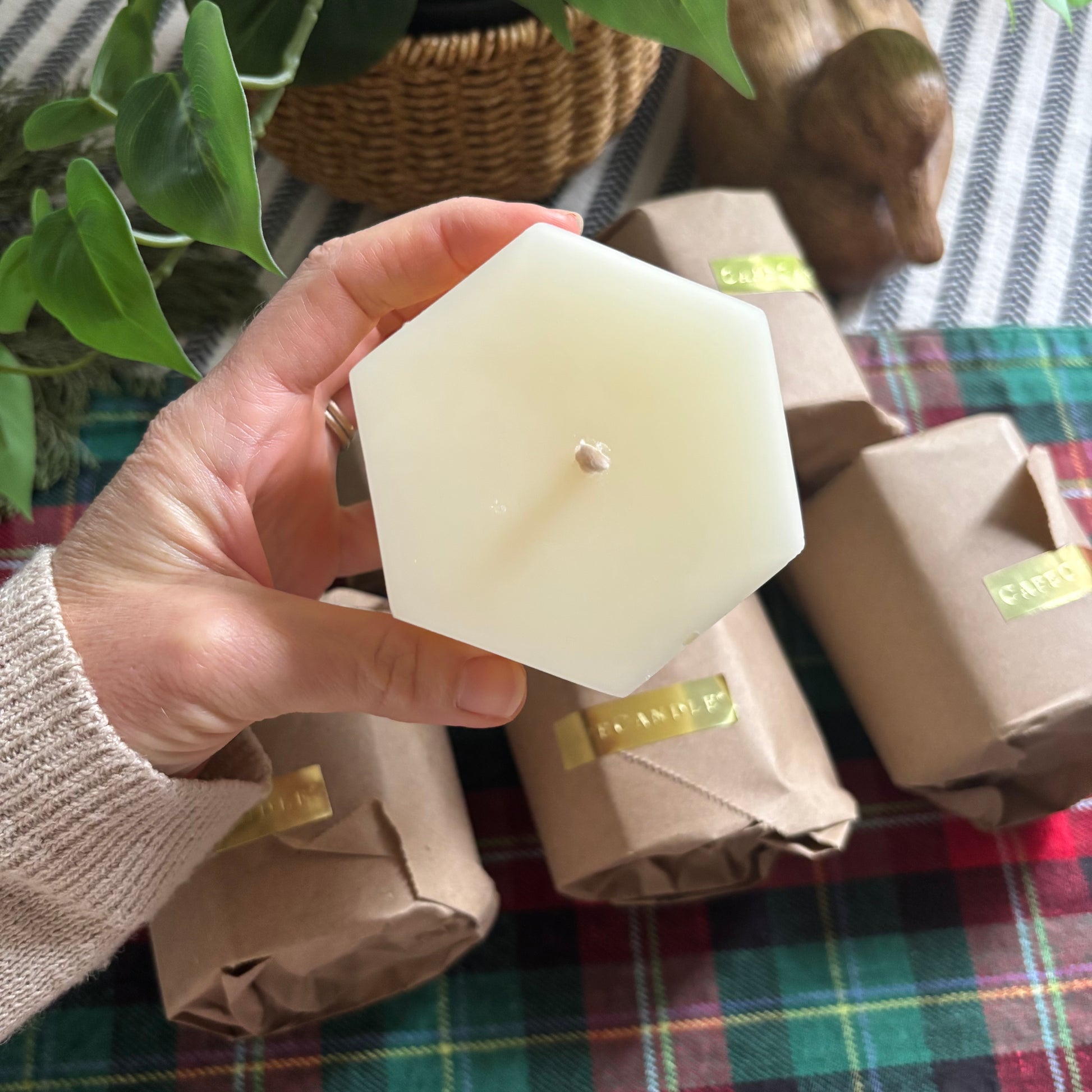 Hand holding a hexagonal white candle with gift-wrapped items and a plaid tablecloth in the background.