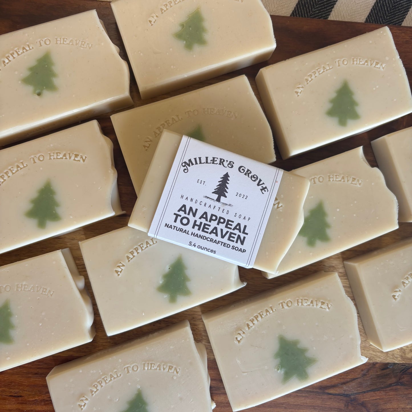 Multiple rectangular bars of handmade soap arranged on a wooden surface, with one bar centered and wrapped in a paper label that reads 'Smiler's Grove Handcrafted Soap An Appeal To Heaven.' Each soap bar is light cream with a small green tree motif and embossed text.