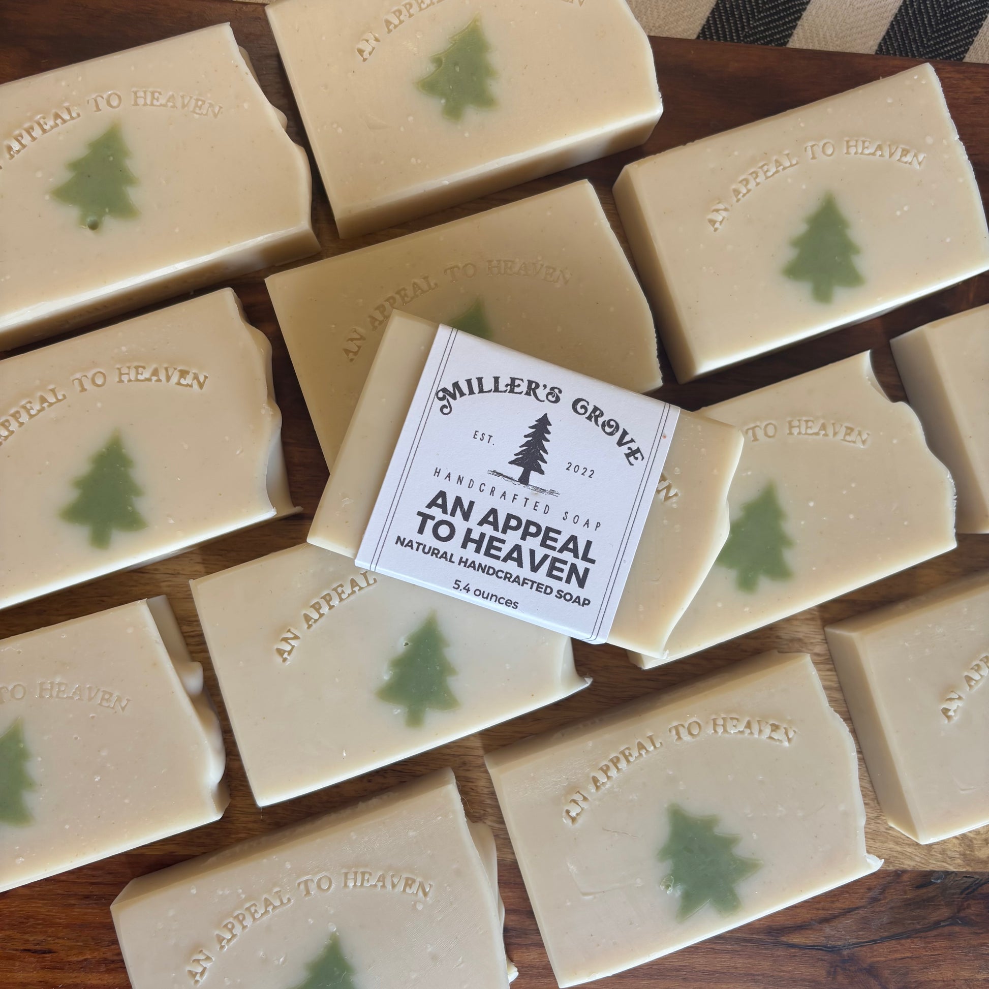 Multiple rectangular bars of handmade soap arranged on a wooden surface, with one bar centered and wrapped in a paper label that reads 'Smiler's Grove Handcrafted Soap An Appeal To Heaven.' Each soap bar is light cream with a small green tree motif and embossed text.