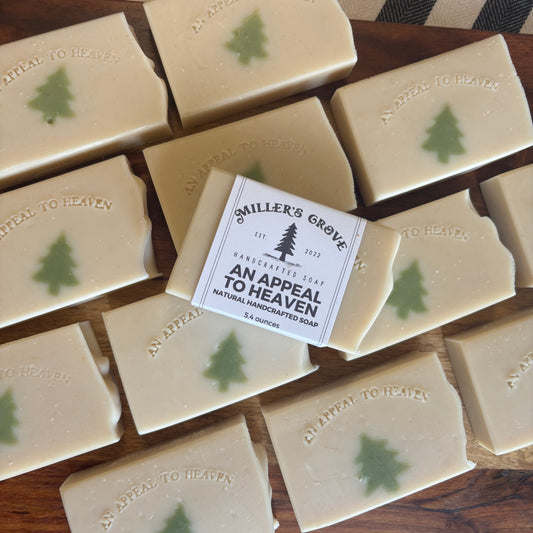 Multiple rectangular bars of handmade soap arranged on a wooden surface, with one bar centered and wrapped in a paper label that reads 'Smiler's Grove Handcrafted Soap An Appeal To Heaven.' Each soap bar is light cream with a small green tree motif and embossed text.