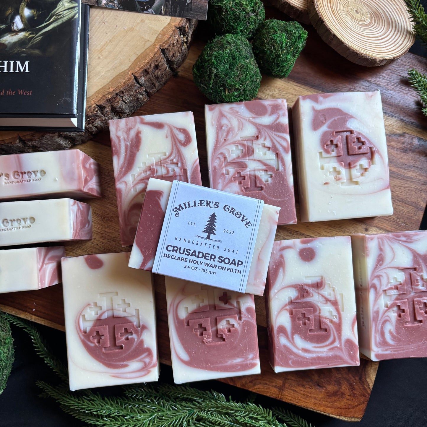 Assorted soap bars with a brand label on a wooden board, surrounded by greenery.