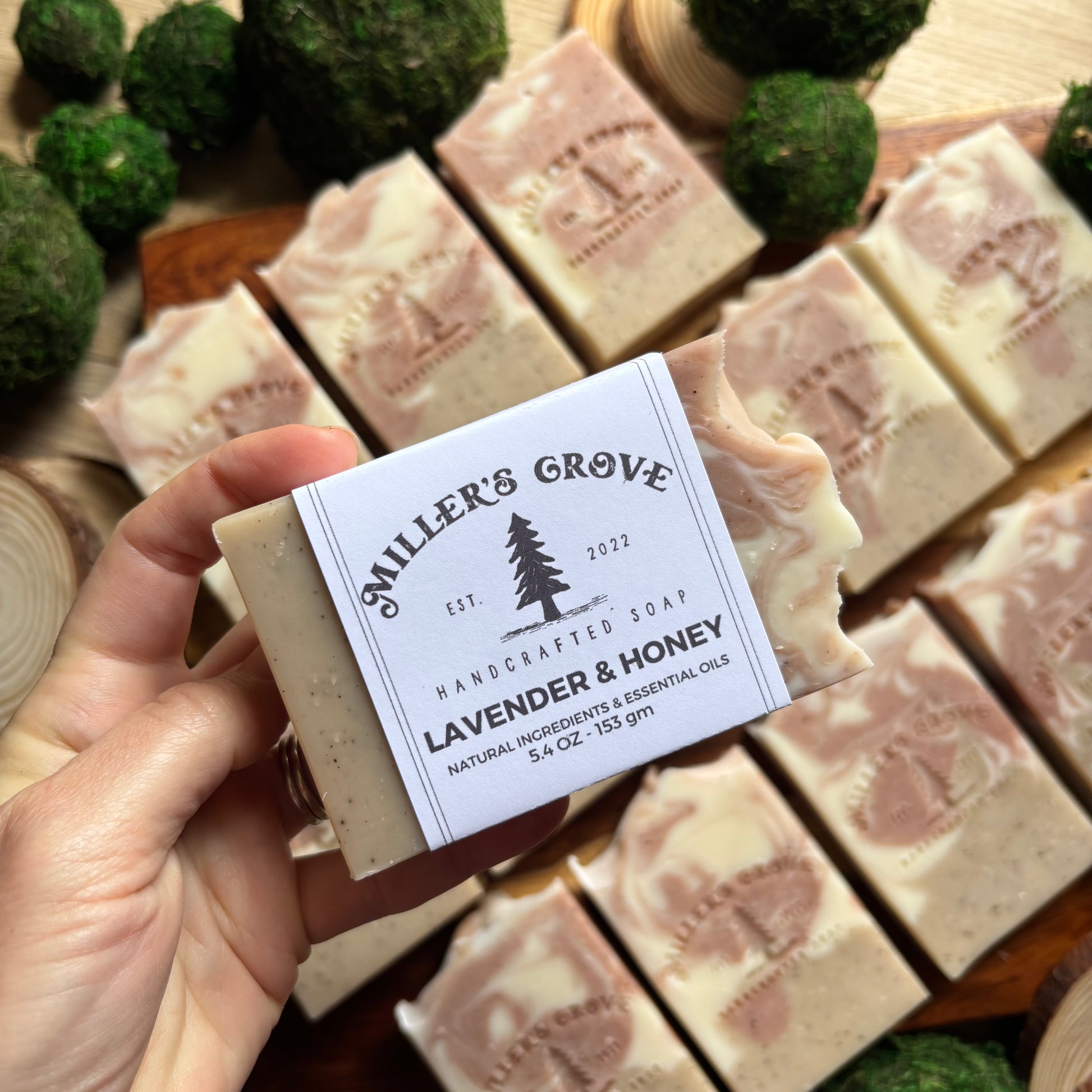 Handcrafted soap bar with 'Miller's Grove' label held by a hand, surrounded by more soap bars.