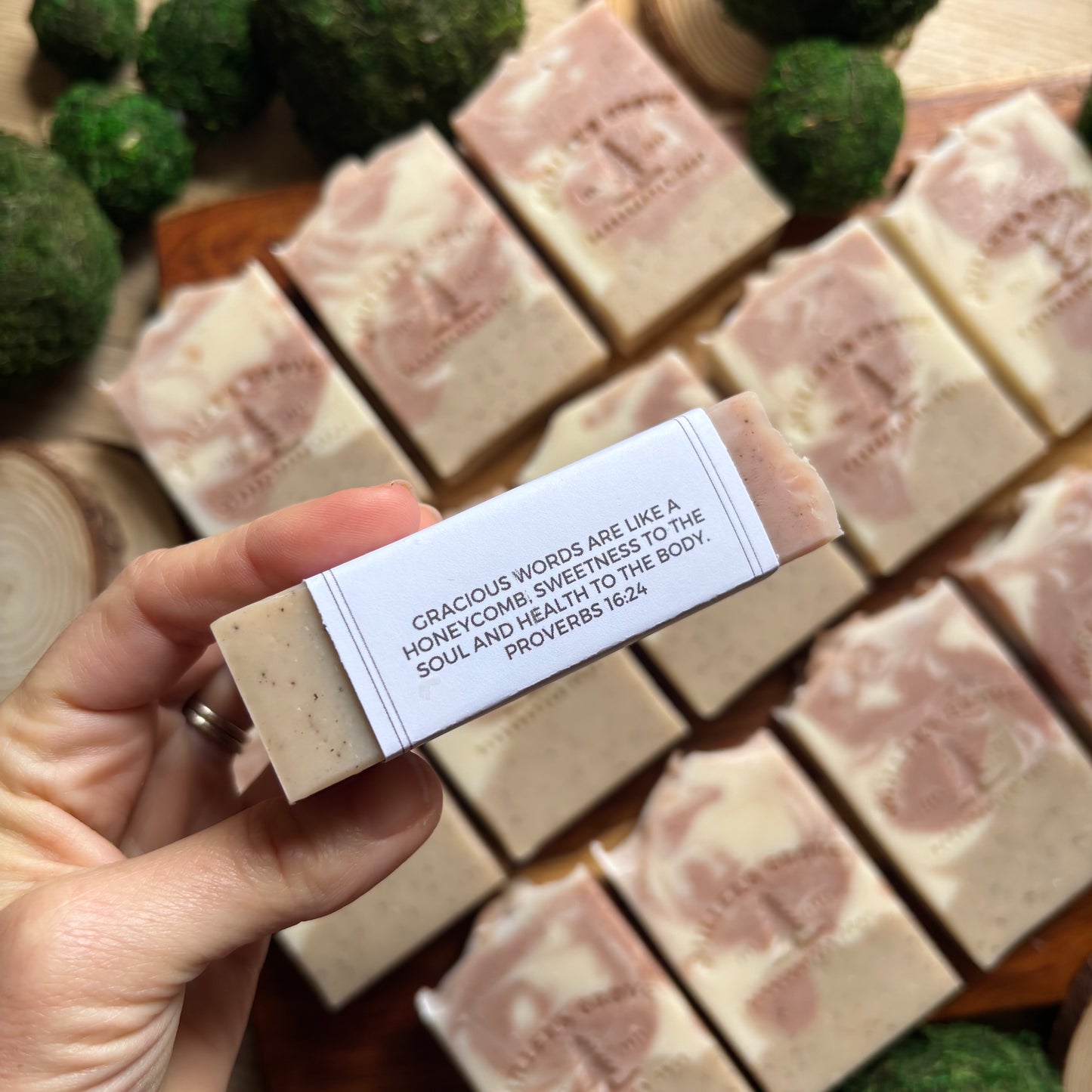 Hand holding a bar of soap with a text label, surrounded by more soap bars and green decorative elements.