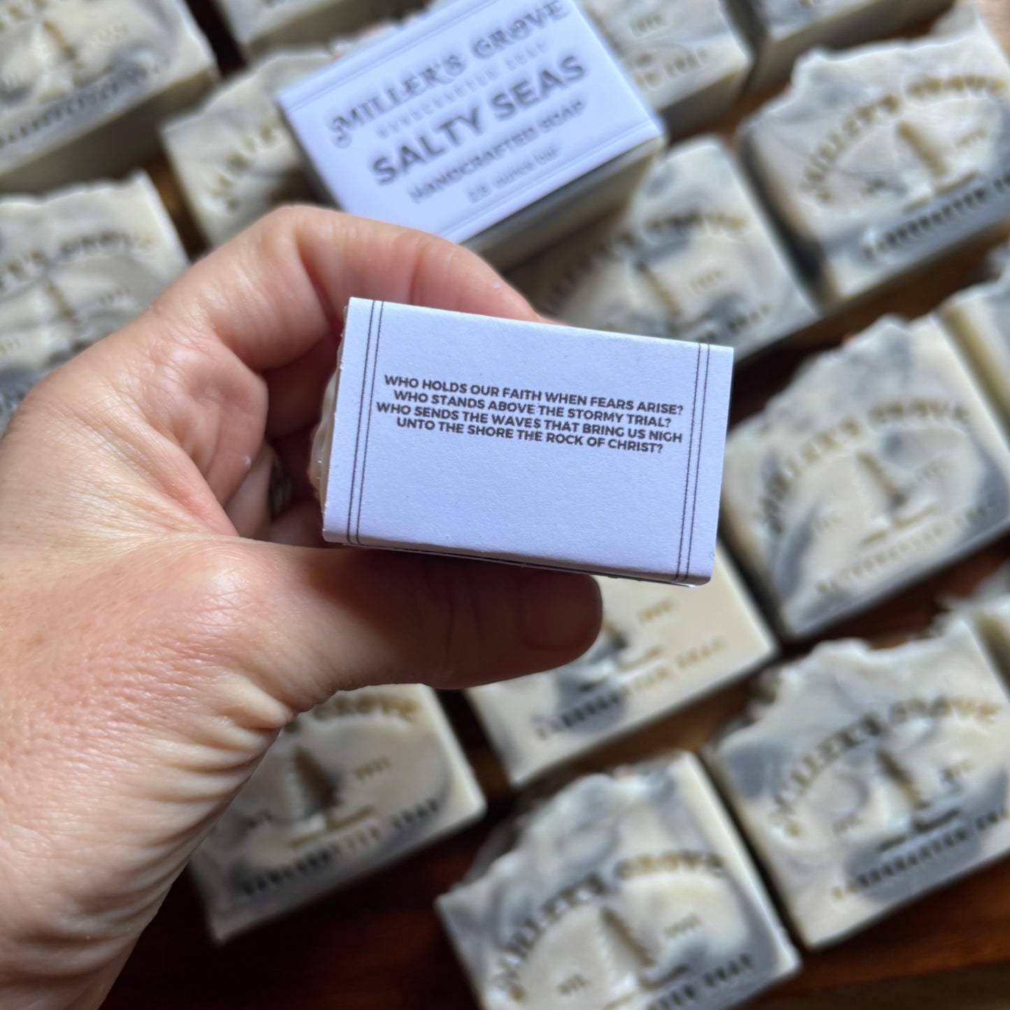 Hand holding a small box of 'Miller's Grove Salty Seas' soap with more soap bars in the background.