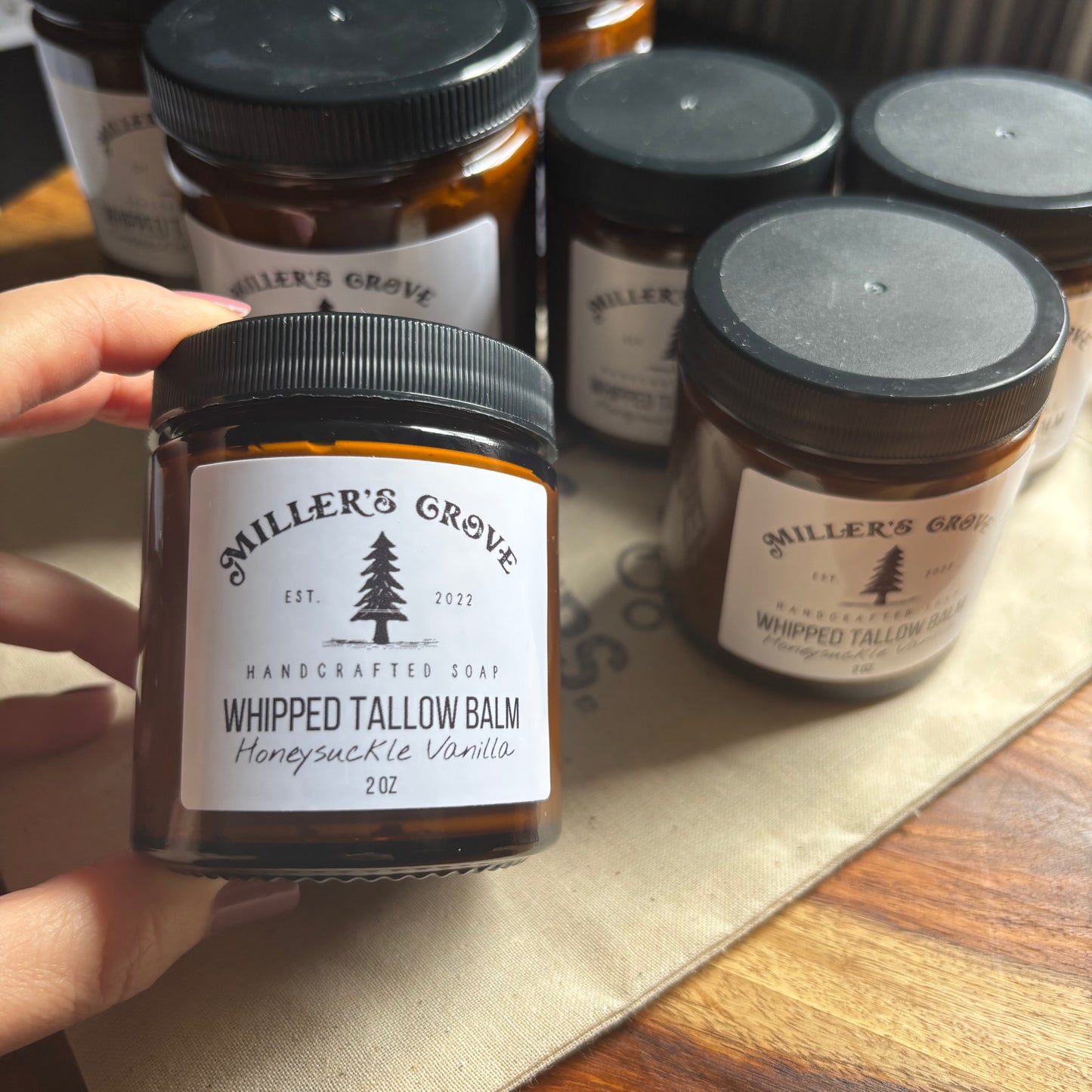 Hand holding a jar of 'Miller's Grove' whipped tallow balm on a wooden surface with more jars in the background.