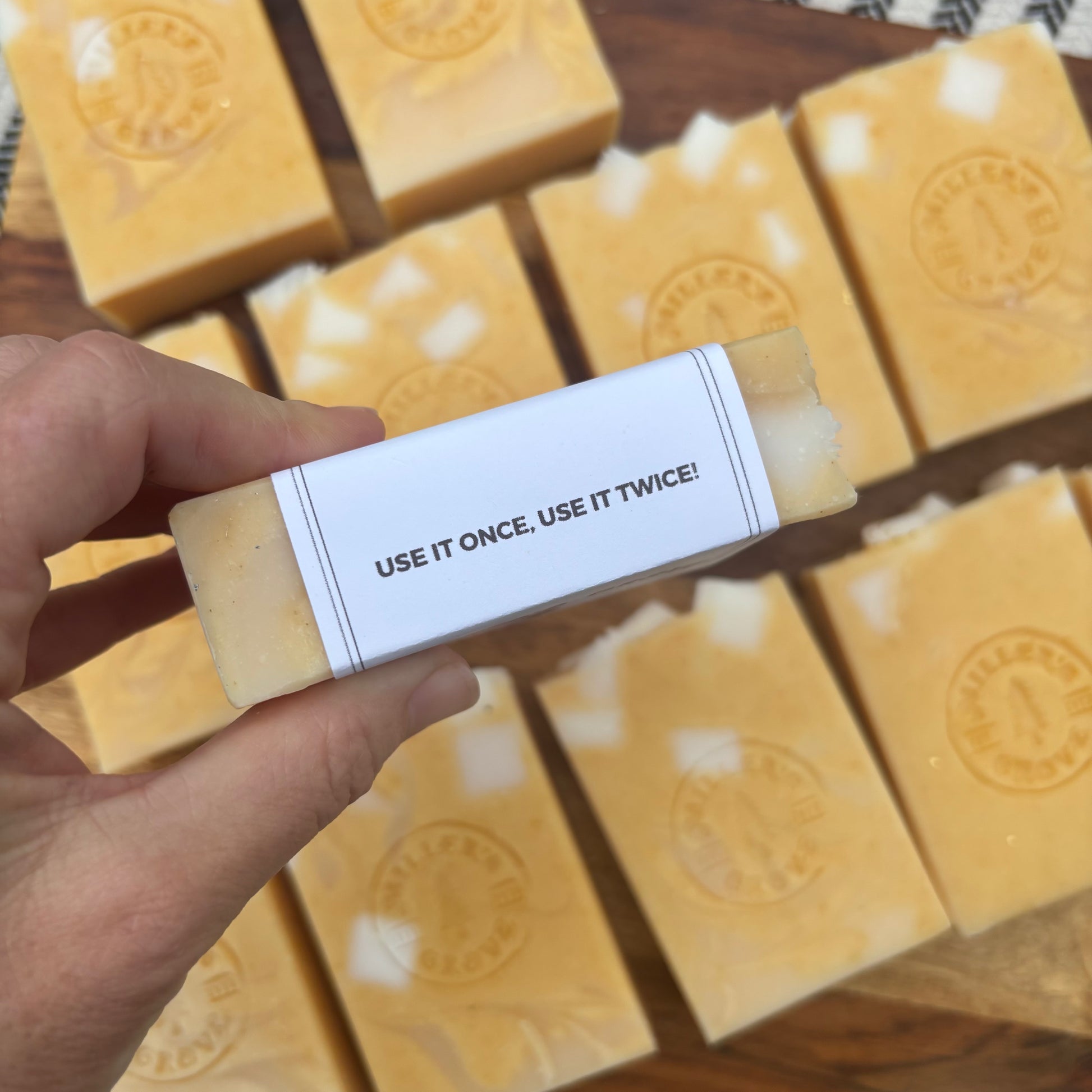 Bar of soap with a label reading 'Use it once, use it twice!' held by a hand, with more bars in the background.