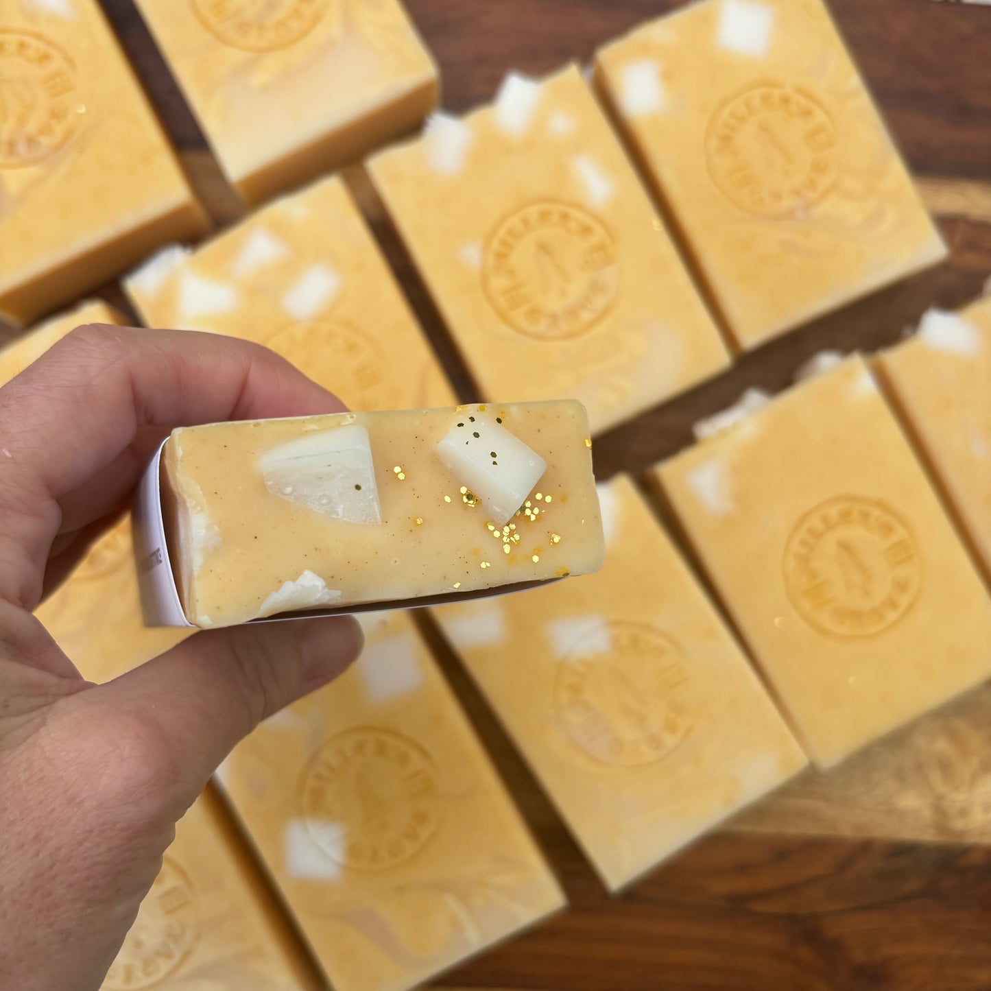 Hand holding a bar of soap with embedded ingredients on a wooden surface with more soap bars.