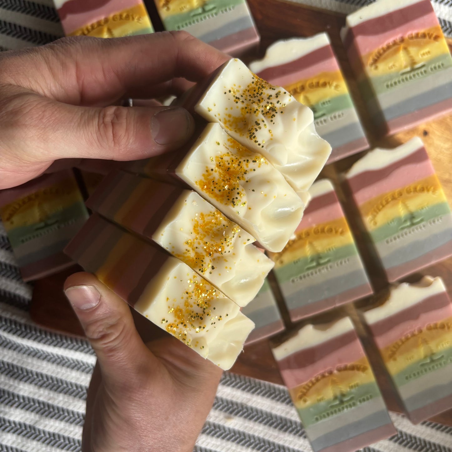 Hand holding a striped soap bar with a wooden tray of more soap bars in the background.