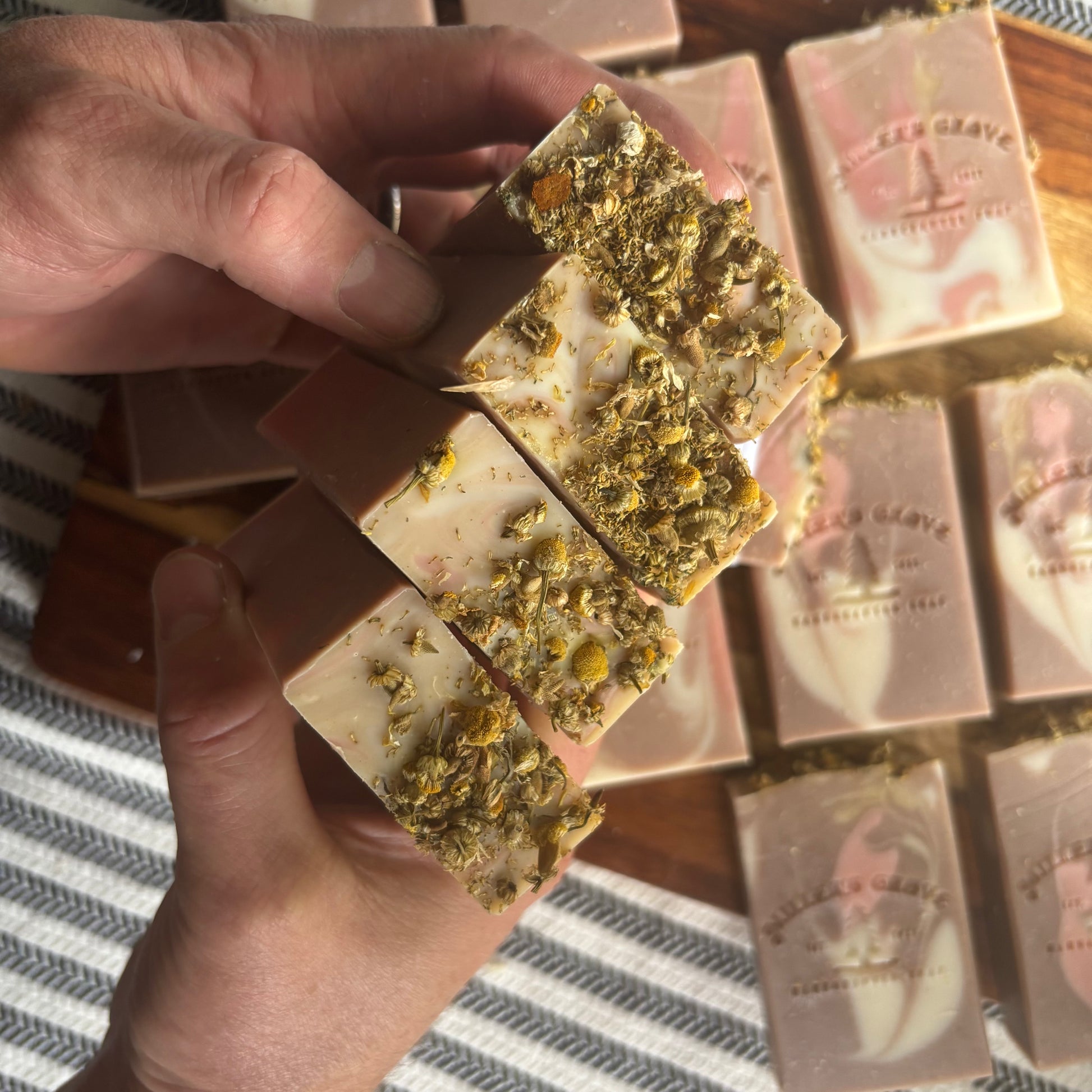 Hand holding a bar of soap with visible herbs, set against a background of similar soap bars.