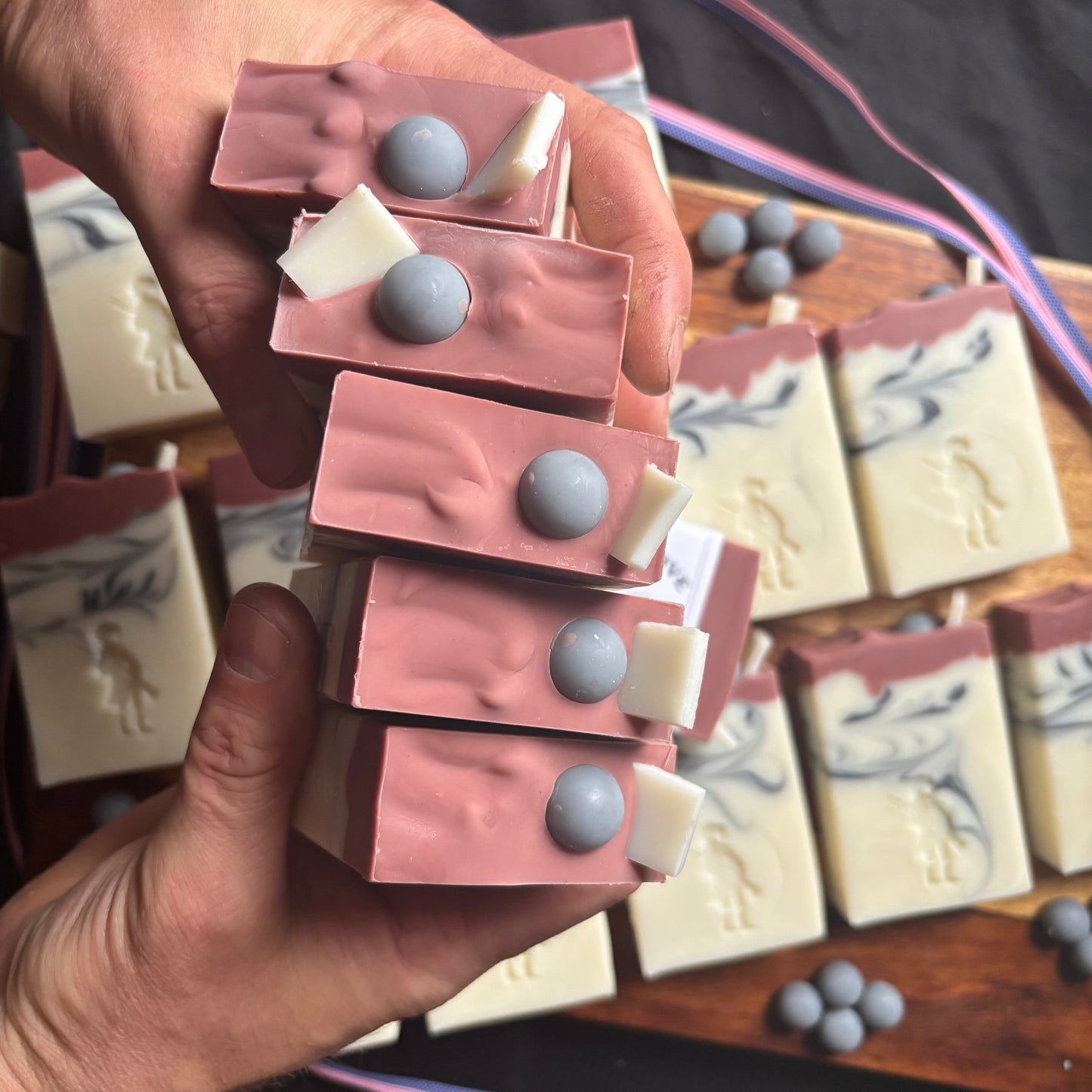 Hand holding a stack of pink and gray soap bars with a blurred background of more soap bars.