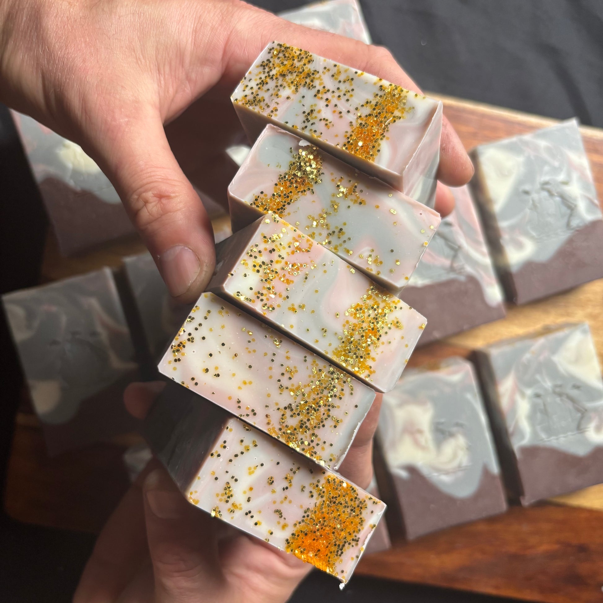 Hand holding a stack of decorative soap bars with glittery toppings on a wooden surface.