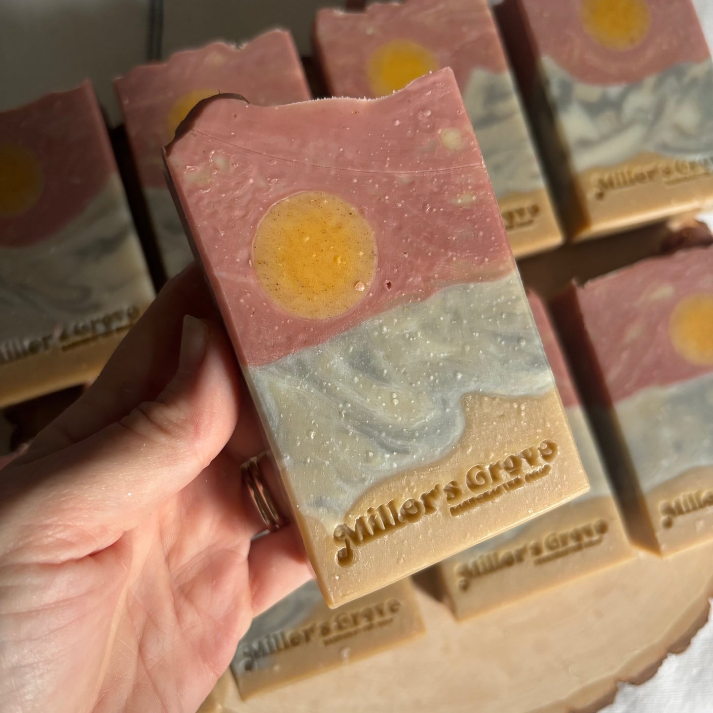 Hand holding a layered soap bar with 'Miller's Grove' branding, surrounded by more soap bars.