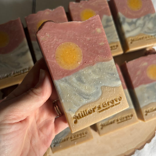 Hand holding a layered soap bar with 'Miller's Grove' branding, surrounded by more soap bars.