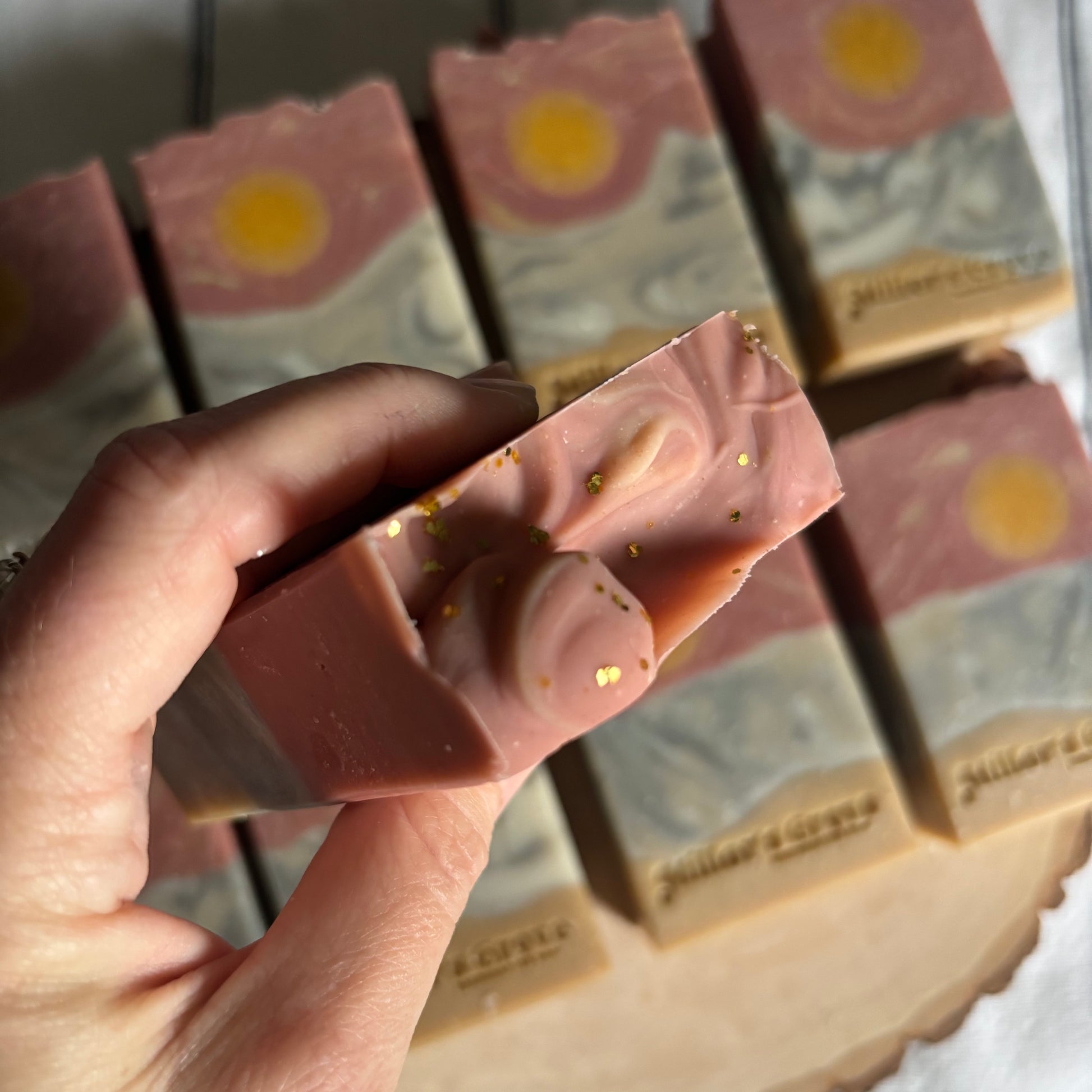 Hand holding a pink soap bar with gold specks, with more soap bars in the background.