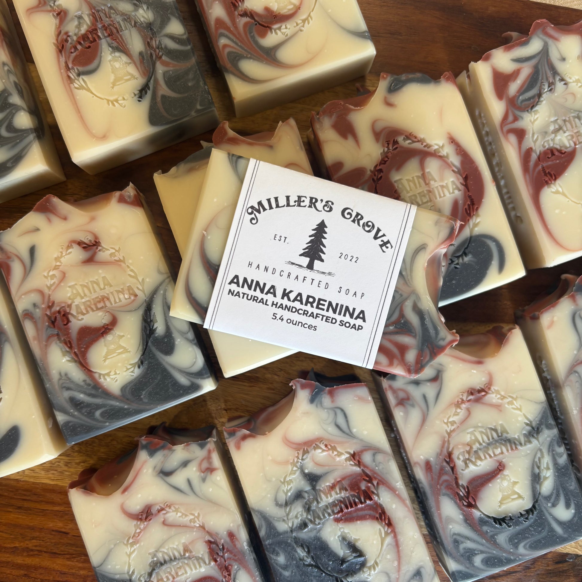 Handcrafted artisan soap bars with cream, red, and dark gray swirls, arranged on a wooden surface. A central bar has a paper label reading 'Miller's Grove Anna Karenina Handcrafted Soap'.