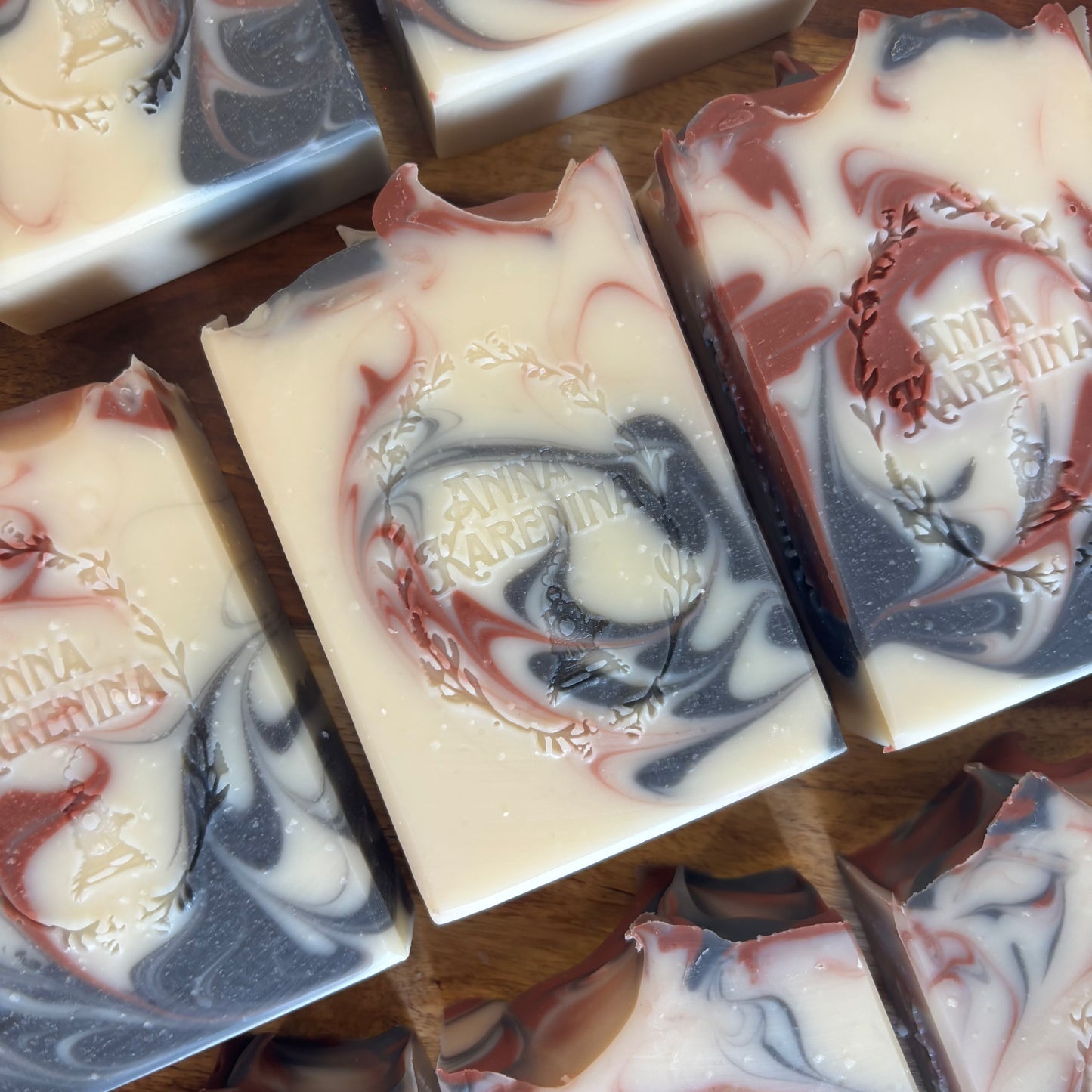Several rectangular artisan soap bars  are arranged on a wooden surface. The soaps have a marbled swirl pattern of creamy off-white, dark charcoal gray, and muted dusty red.
