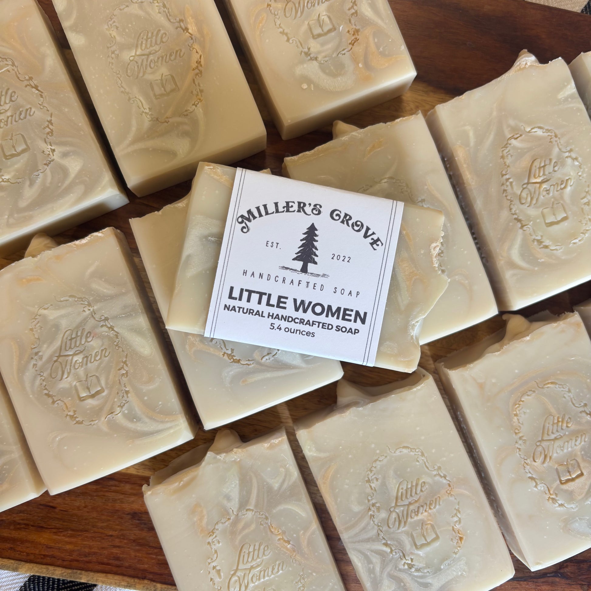 A stack of handmade soap bars on a wooden surface, with the center bar labeled Miller's Grove Little Women Handcrafted Soap.