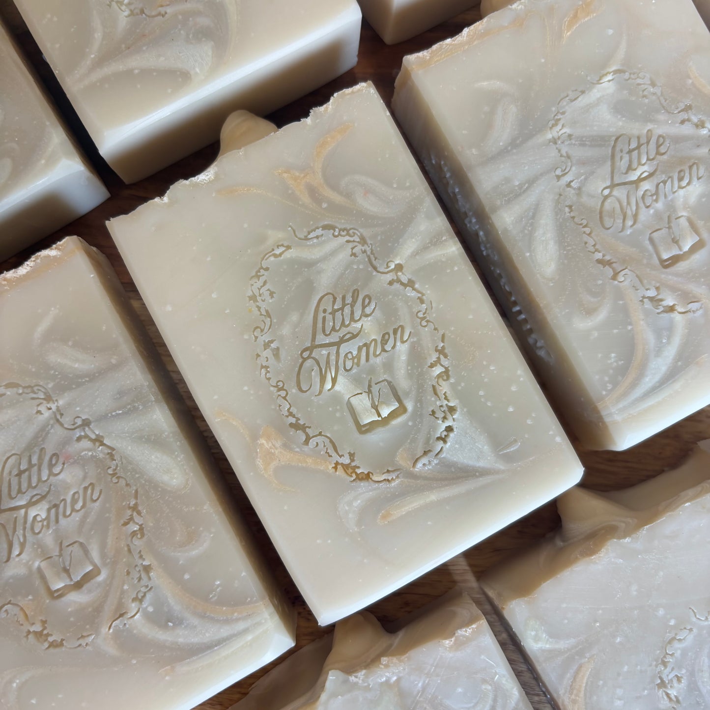 Several rectangular handmade soap bars with  are arranged on a wooden surface. The soaps are off-white/cream with subtle marbled swirls and a small embossed logo reading 'Little Women' in the center.