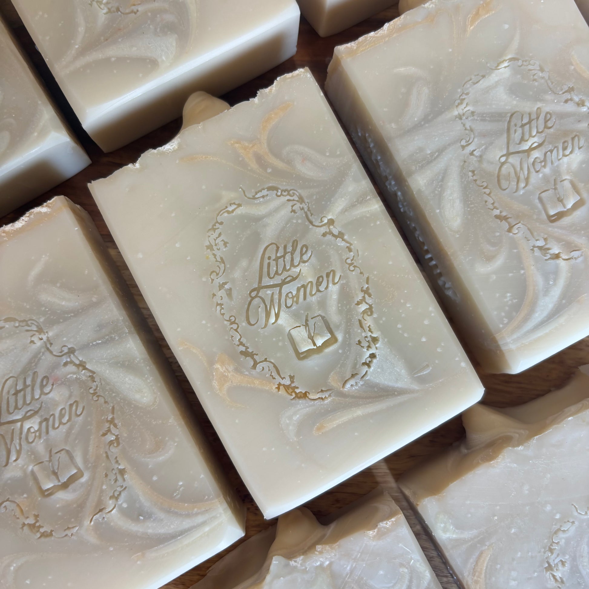 Several rectangular handmade soap bars with  are arranged on a wooden surface. The soaps are off-white/cream with subtle marbled swirls and a small embossed logo reading 'Little Women' in the center.