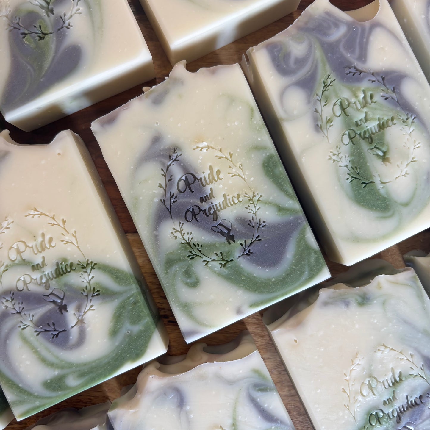 Several handmade soap bars with swirled green, lavender, and cream patterns, each embossed with the brand name 'Pride and Prejudice", arranged on a wooden surface.