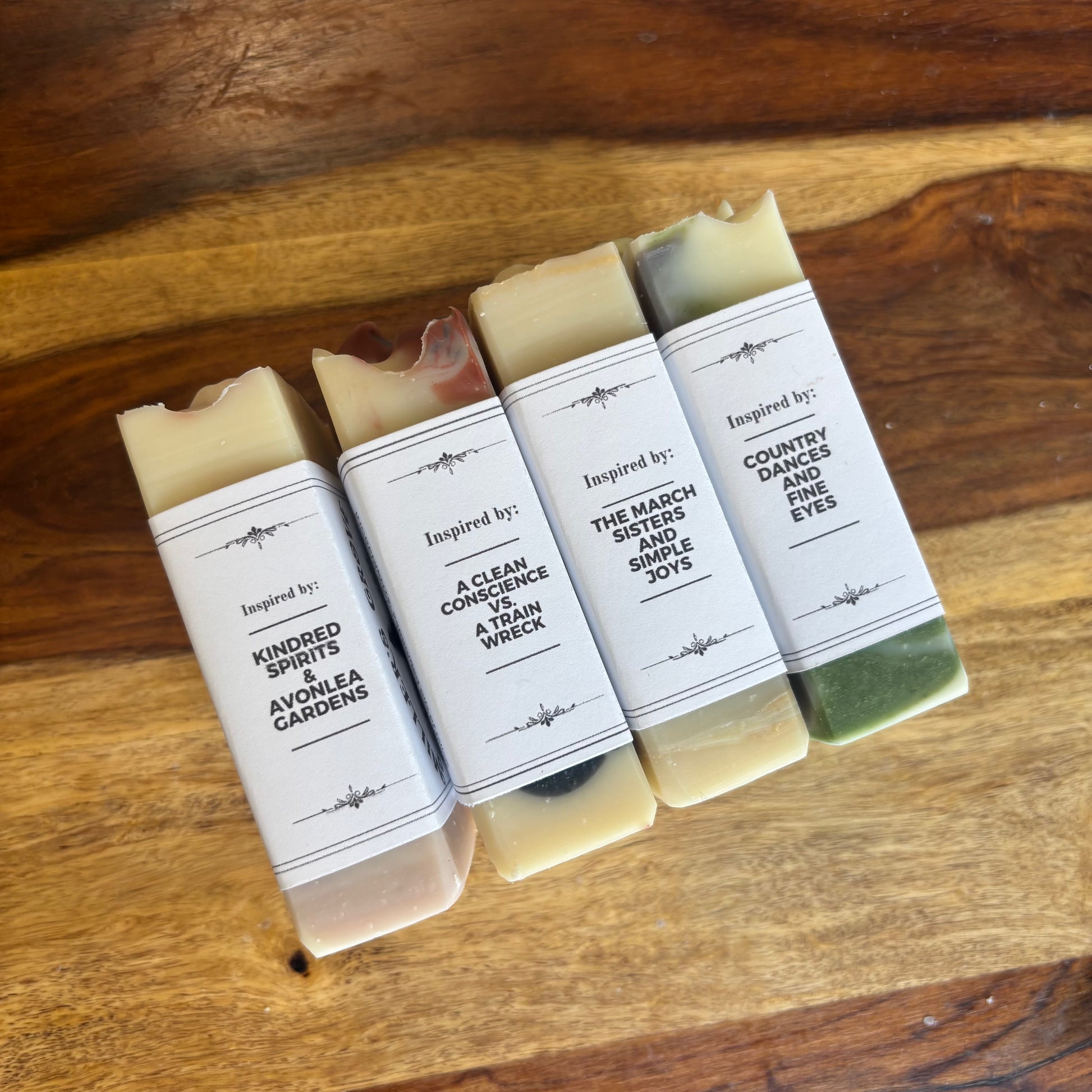 Four rectangular bars of handmade soap arranged on a wooden surface, each wrapped with a white paper label that reads 'Inspired by: [different phrases]'. The soaps show natural marbled colors including beige, green, and brown.