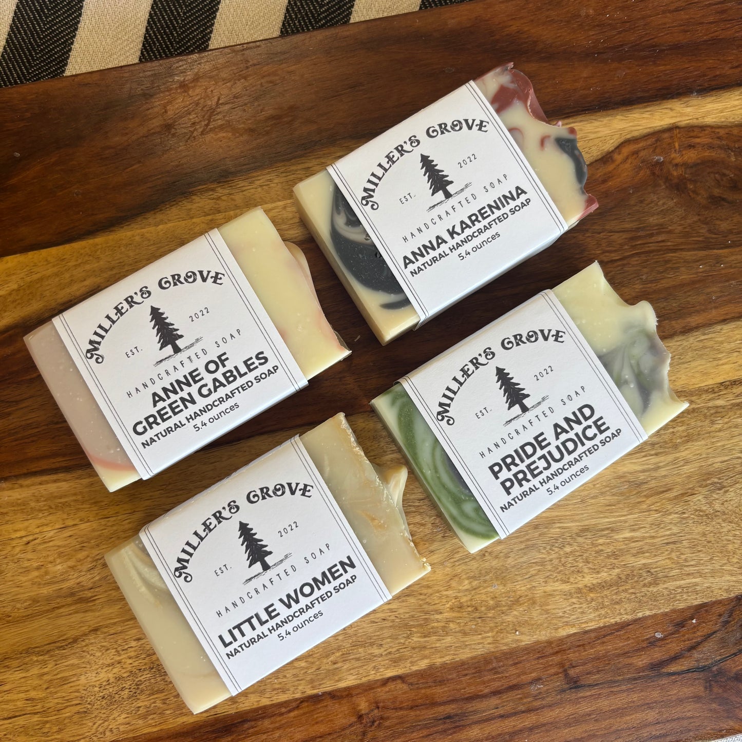 Four bars of handmade soap from Miller's Grove arranged on a wooden surface. The soaps include Anne of Green Cables, Anna Karenina, Little Women, and Pride and Prejudice, each with a white paper label featuring a black tree logo.