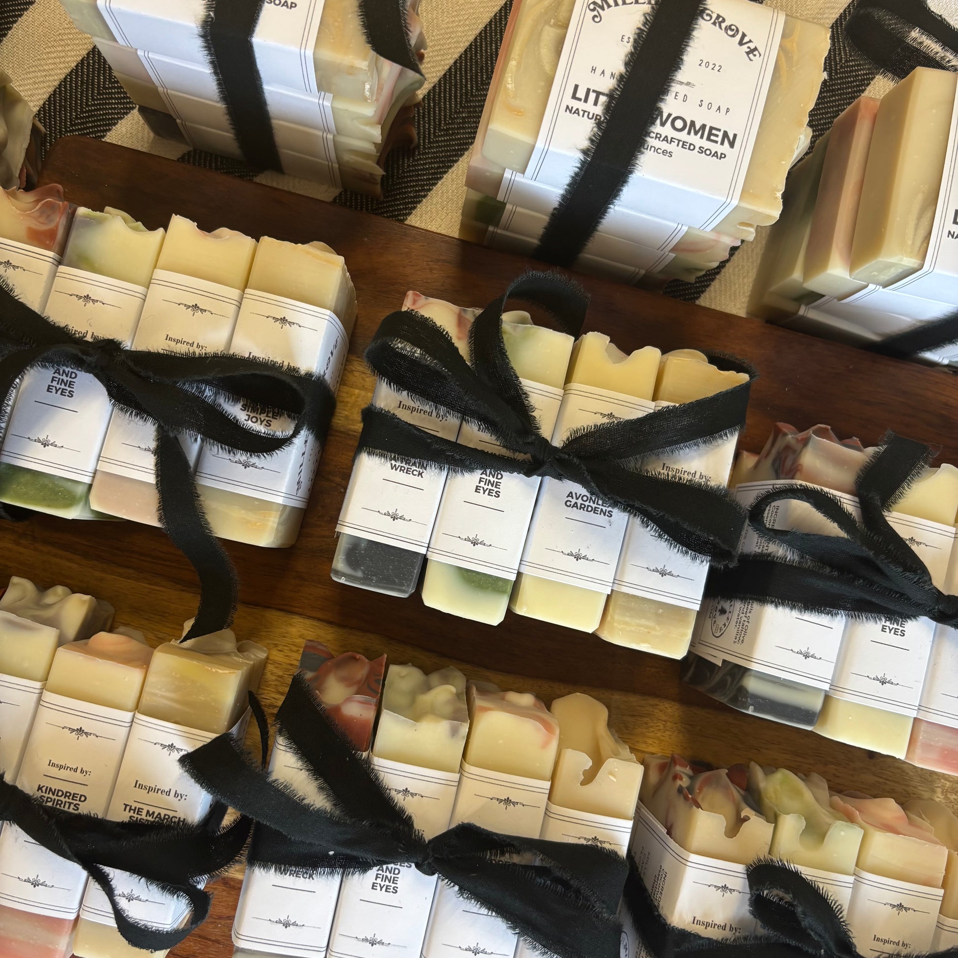 Multiple wrapped bars of handmade soap arranged on wooden trays, tied with black ribbon and labeled with white paper tags. The soap bars show natural cream, yellow, pink, and green tones.