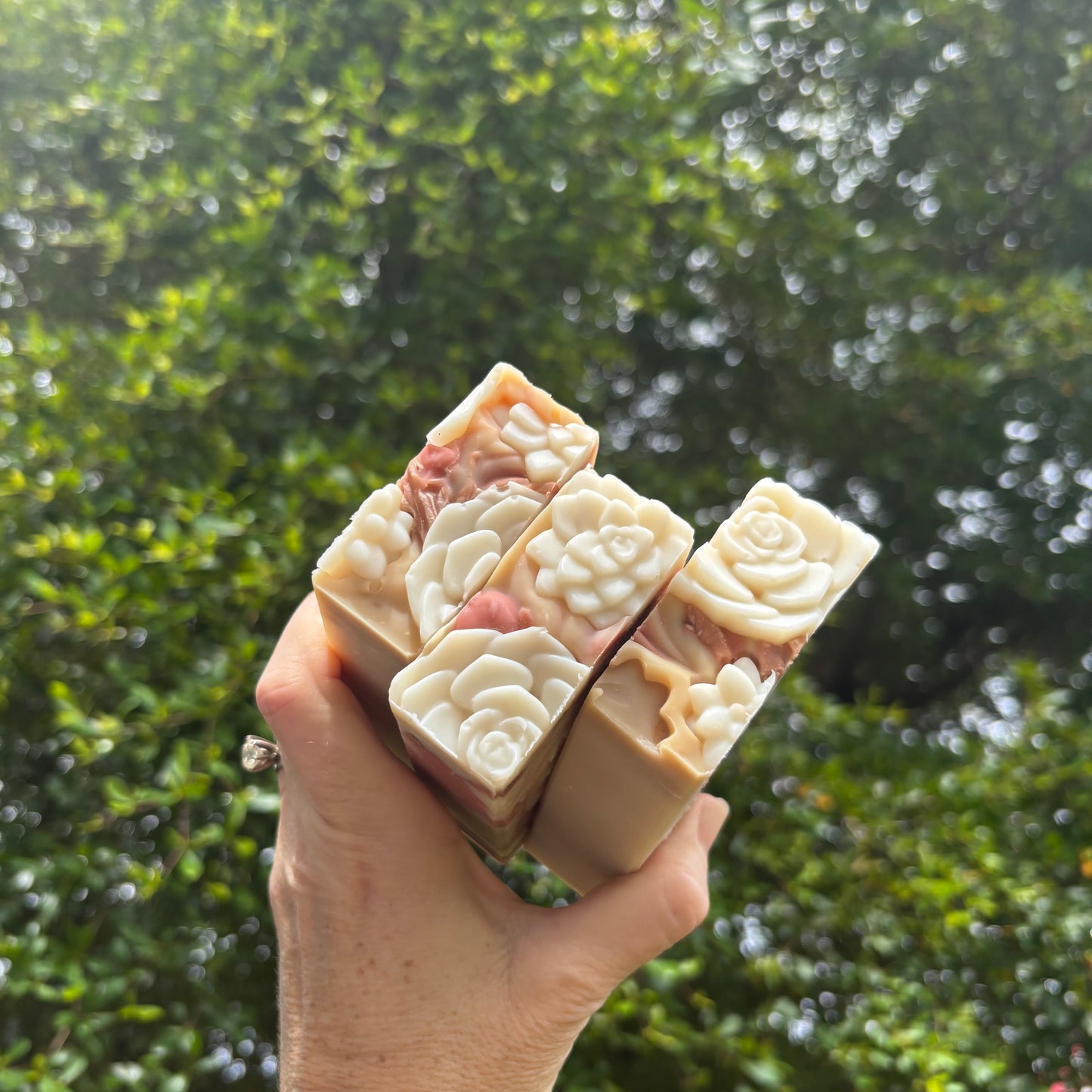 Three decorative handmade soap bars with embossed floral patterns, held in a hand against a blurred background of green foliage.