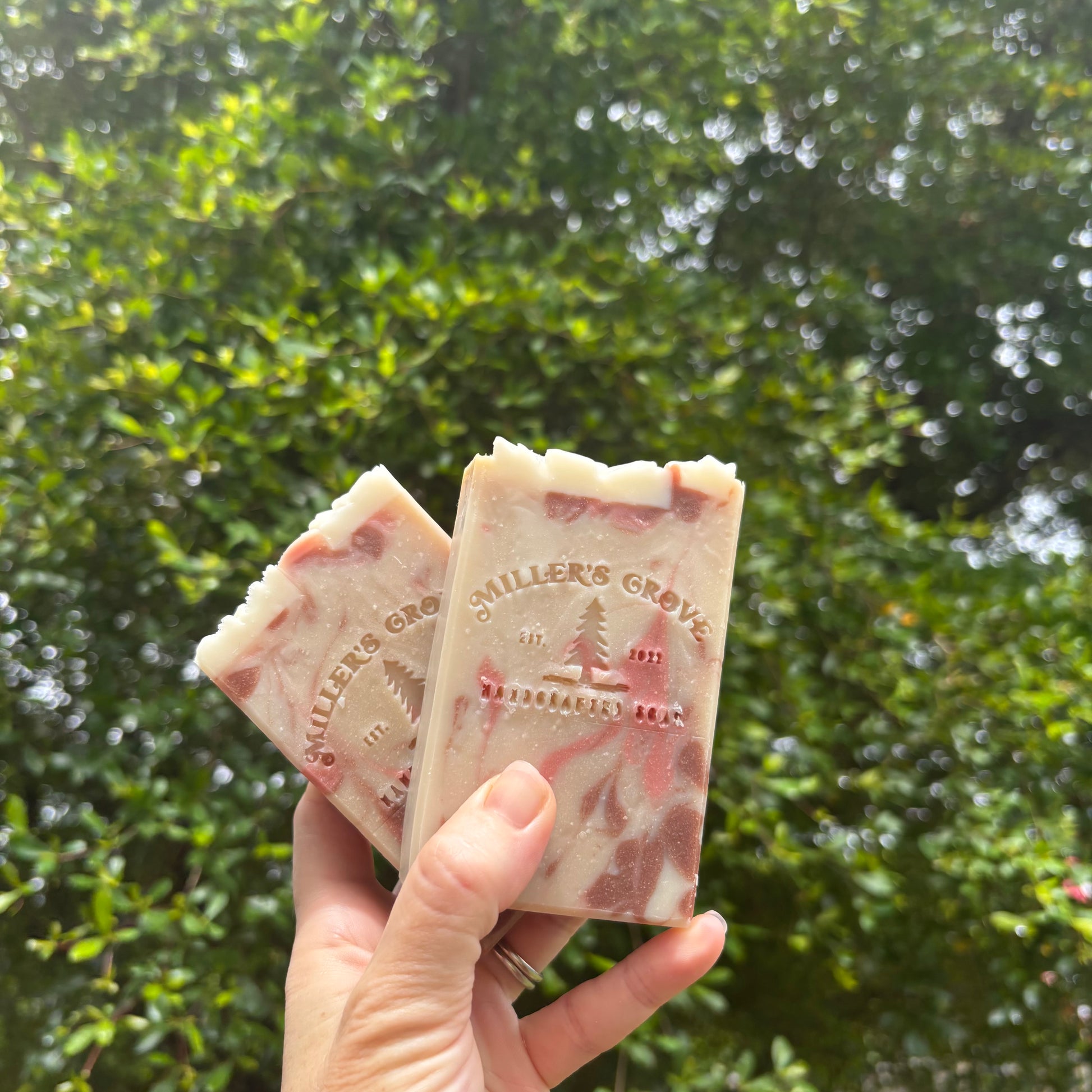 A hand holds two handmade soap bars with a pink and cream marbled pattern, embossed with the brand name Miller's Grove', against a blurred background of green foliage.