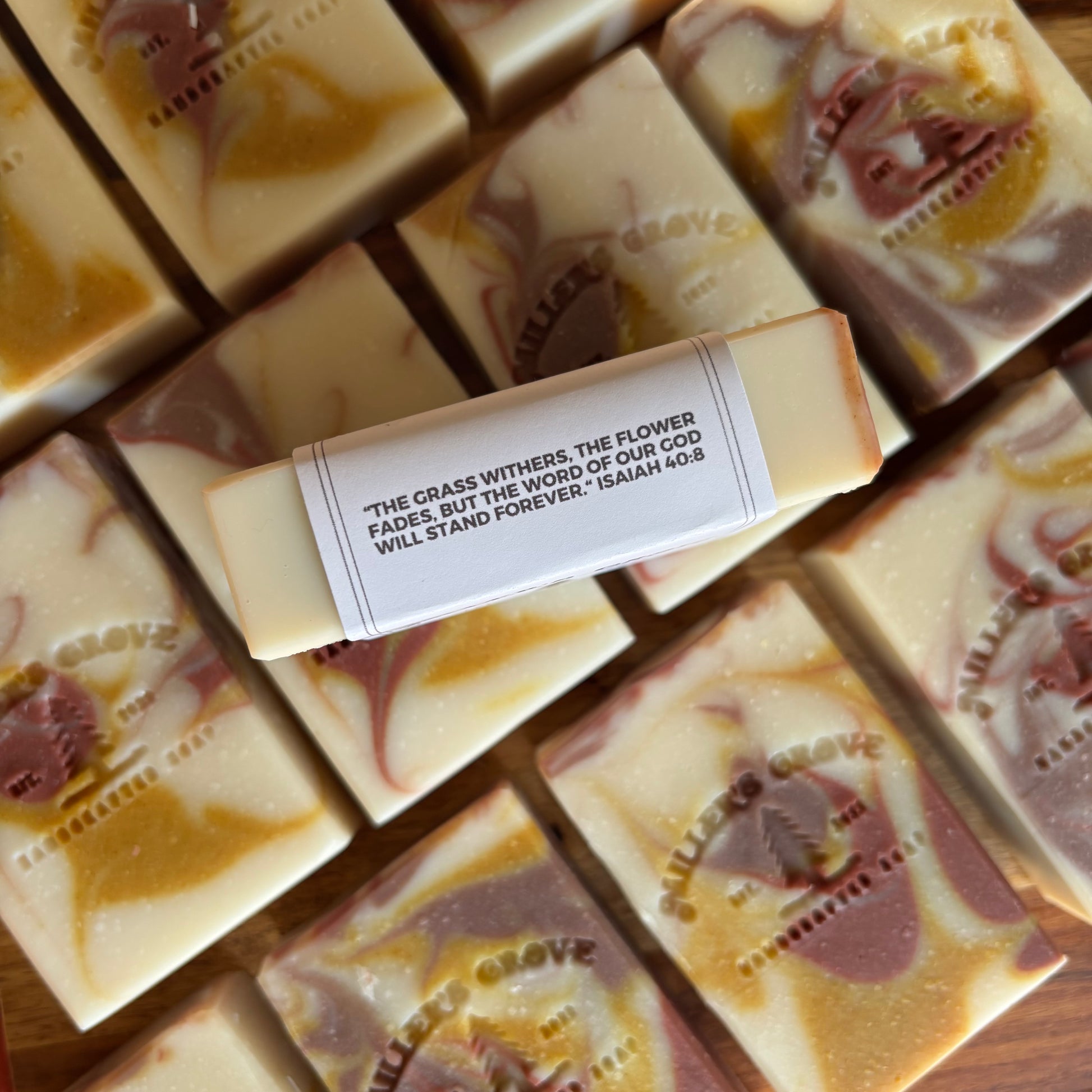 A close-up of several handmade soap bars arranged on a wooden surface, with one soap bar centered and wrapped in a white paper band that reads, 'THE GRASS WITHERS, THE FLOWER FADES, BUT THE WORD OF OUR GOD WILL STAND FOREVER; ISAIAH 40:8.' The soaps have a marbled cream, tan, and muted mauve-brown color pattern, and the brand name 'Millers GROVE' is embossed on the soaps.