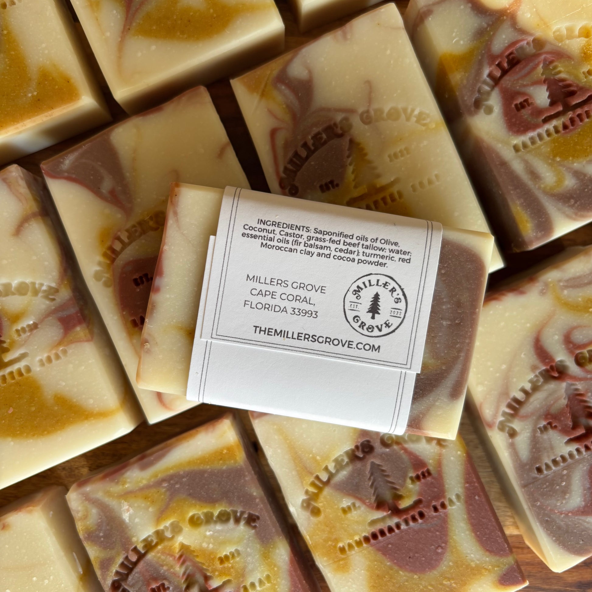A top-down close-up of handmade soap bars arranged closely together, with a white paper label wrapped around one bar. The soaps have a creamy off-white base with swirls of muted pink, tan, and golden yellow, and the brand name MILLERS GROVE is embossed on the surface.