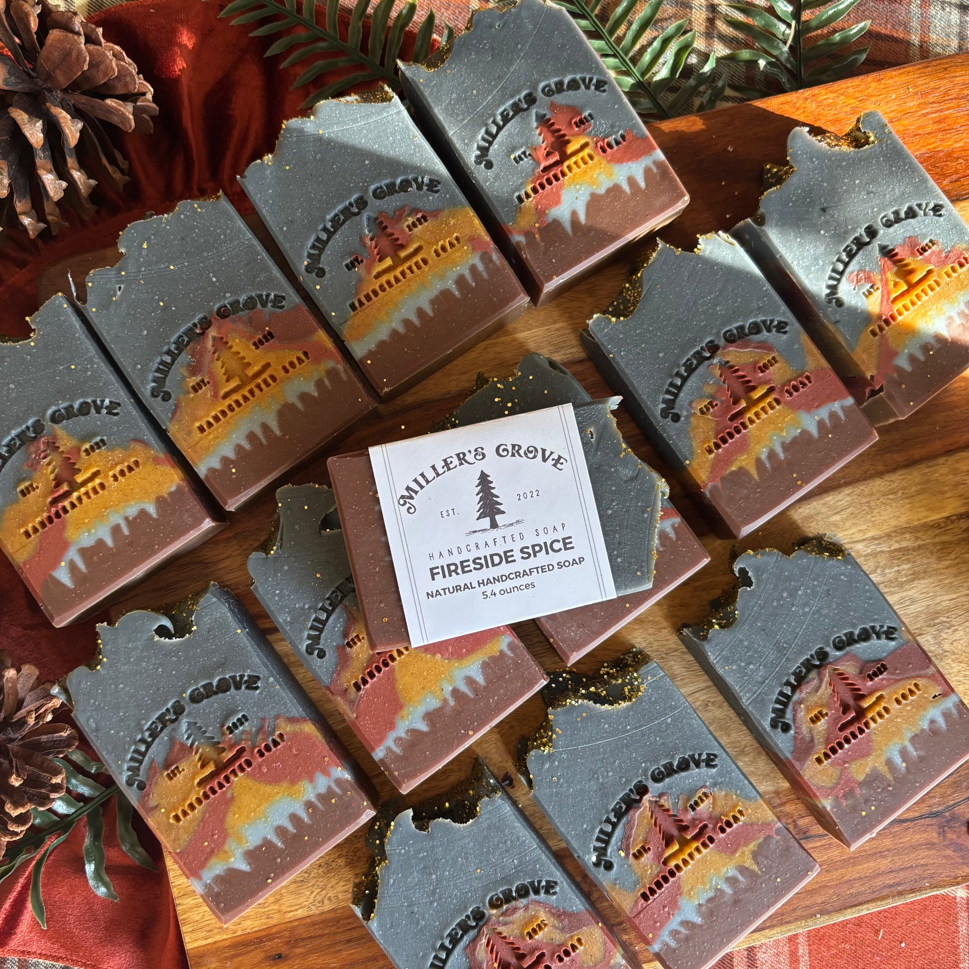 Multiple bars of Miller's Grove Handcrafted Soap in the 'Fireside Spice' scent are arranged on a wooden surface with pinecones and greenery. The soap bars are dark gray with a printed label showing a cabin scene in orange, yellow, and red, with a central white paper label.