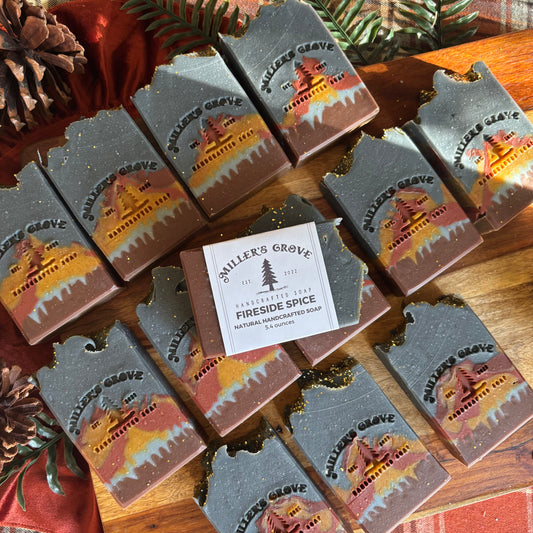 Multiple bars of Miller's Grove Handcrafted Soap in the 'Fireside Spice' scent are arranged on a wooden surface with pinecones and greenery. The soap bars are dark gray with a printed label showing a cabin scene in orange, yellow, and red, with a central white paper label.