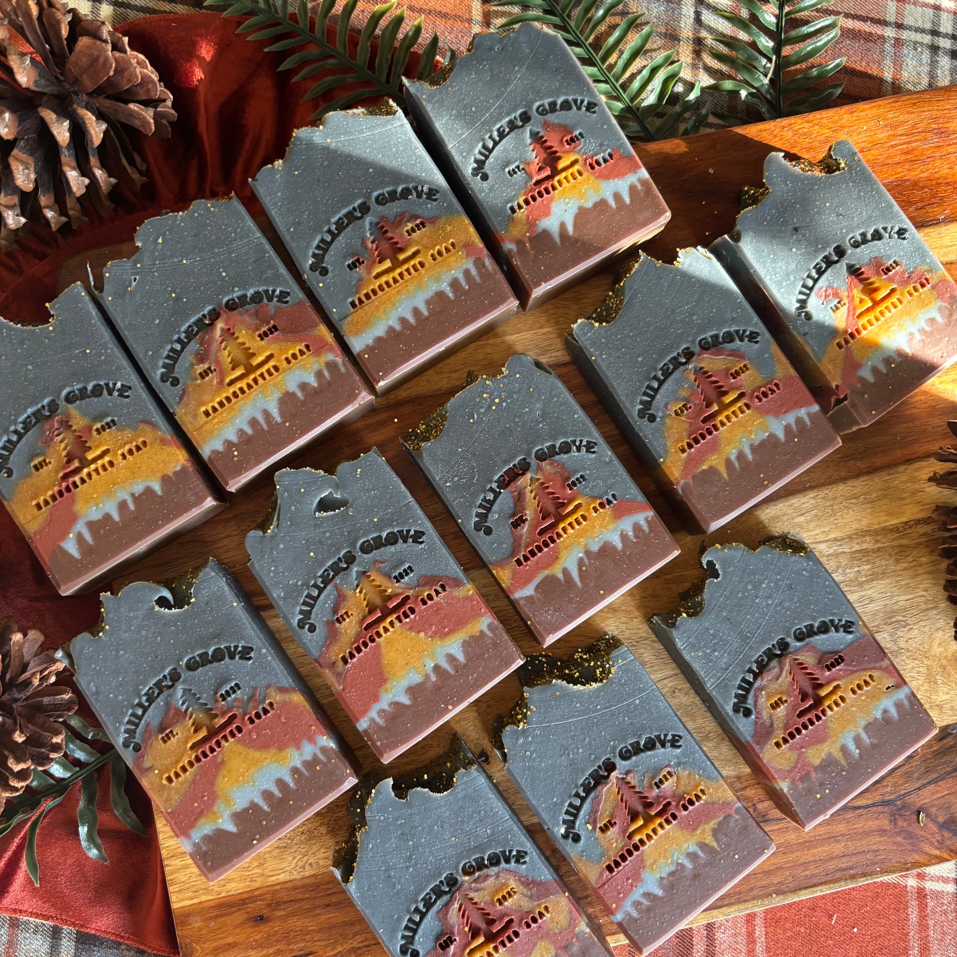 A set of handmade soap bars with rustic, jagged edges, decorated with a scenic 'Miller's Grove' tree illustration and gold glitter accents, arranged on a wooden surface with pinecones and holiday greenery.