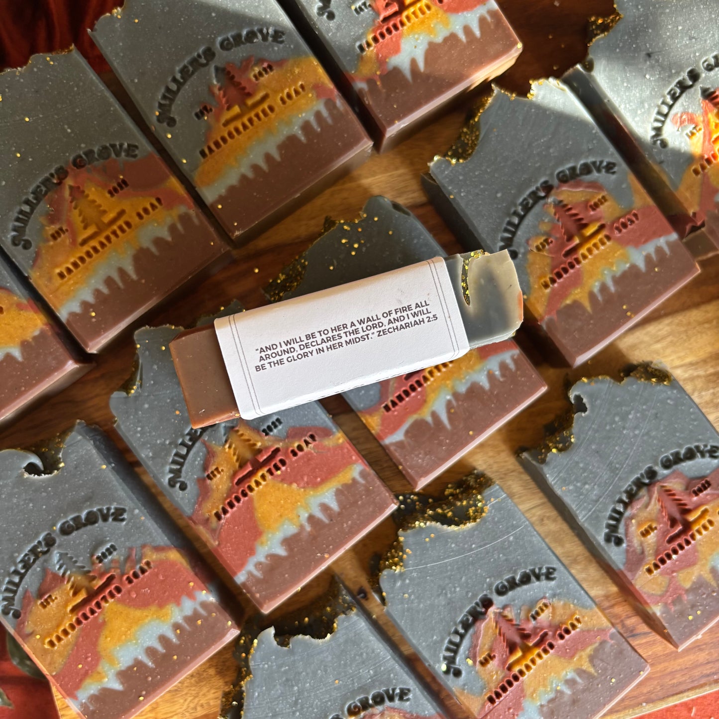 Multiple rectangular artisan soaps arranged on a wooden surface, each printed with the words "Miller's Grove" and a scenic cemetery landscape design in orange, yellow, and red tones. One soap is broken open to reveal the solid bar inside, with a small paper label attached.