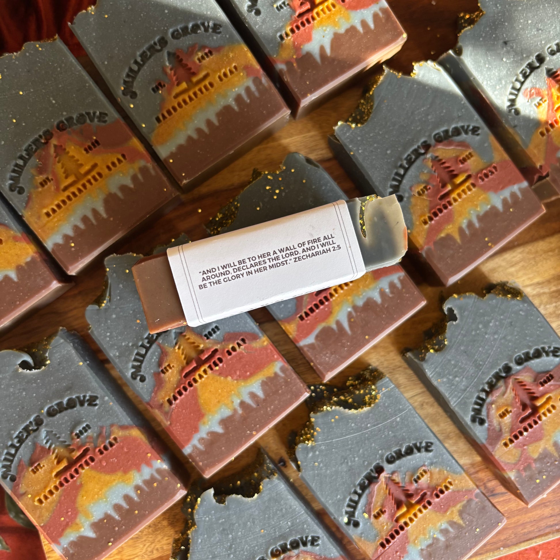 Multiple rectangular artisan soaps arranged on a wooden surface, each printed with the words "Miller's Grove" and a scenic cemetery landscape design in orange, yellow, and red tones. One soap is broken open to reveal the solid bar inside, with a small paper label attached.