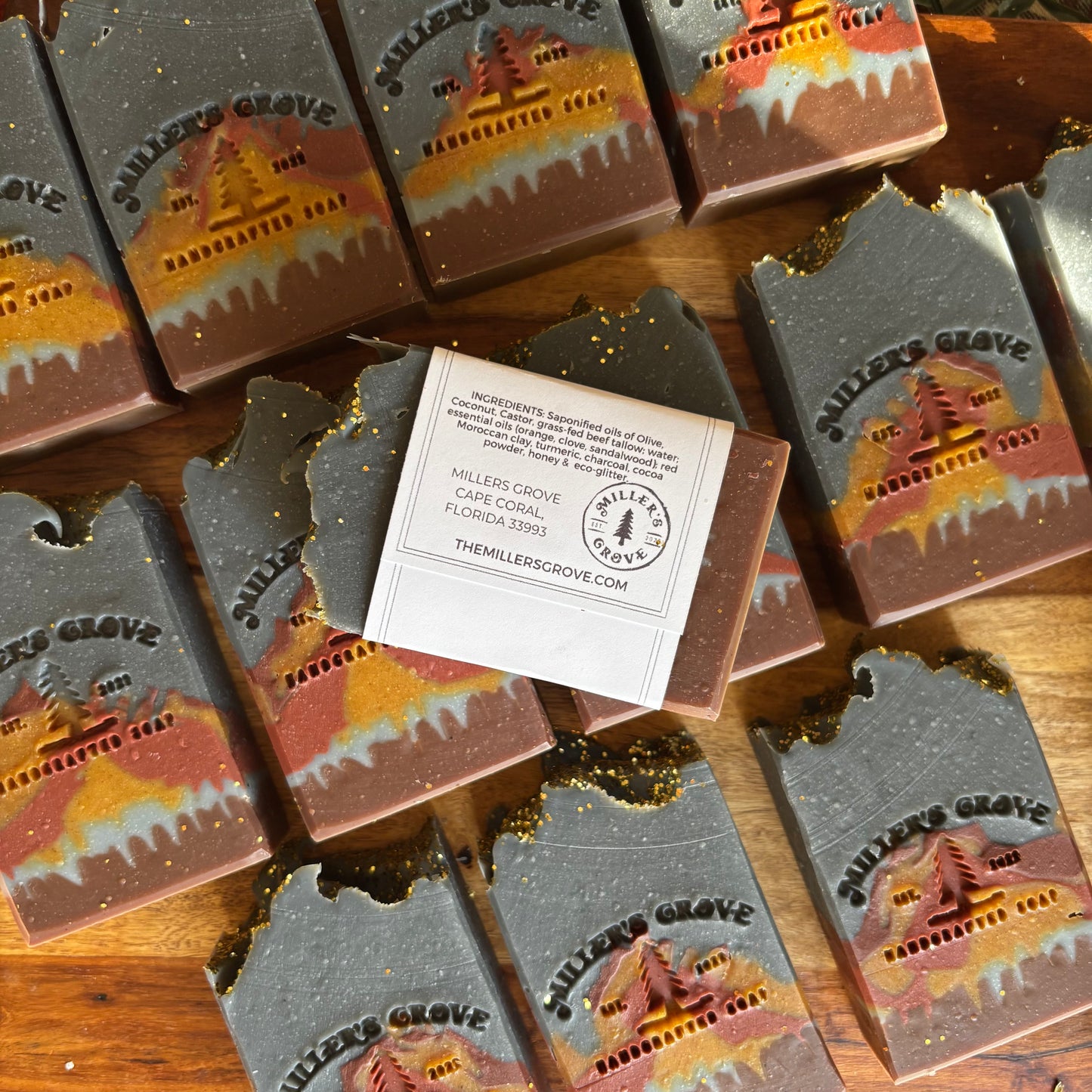 Handcrafted artisan soap bars from Millers Grove, Florida, featuring a scenic mountain cabin design with gold glitter accents, arranged on a wooden surface with one bar wrapped in a paper band.
