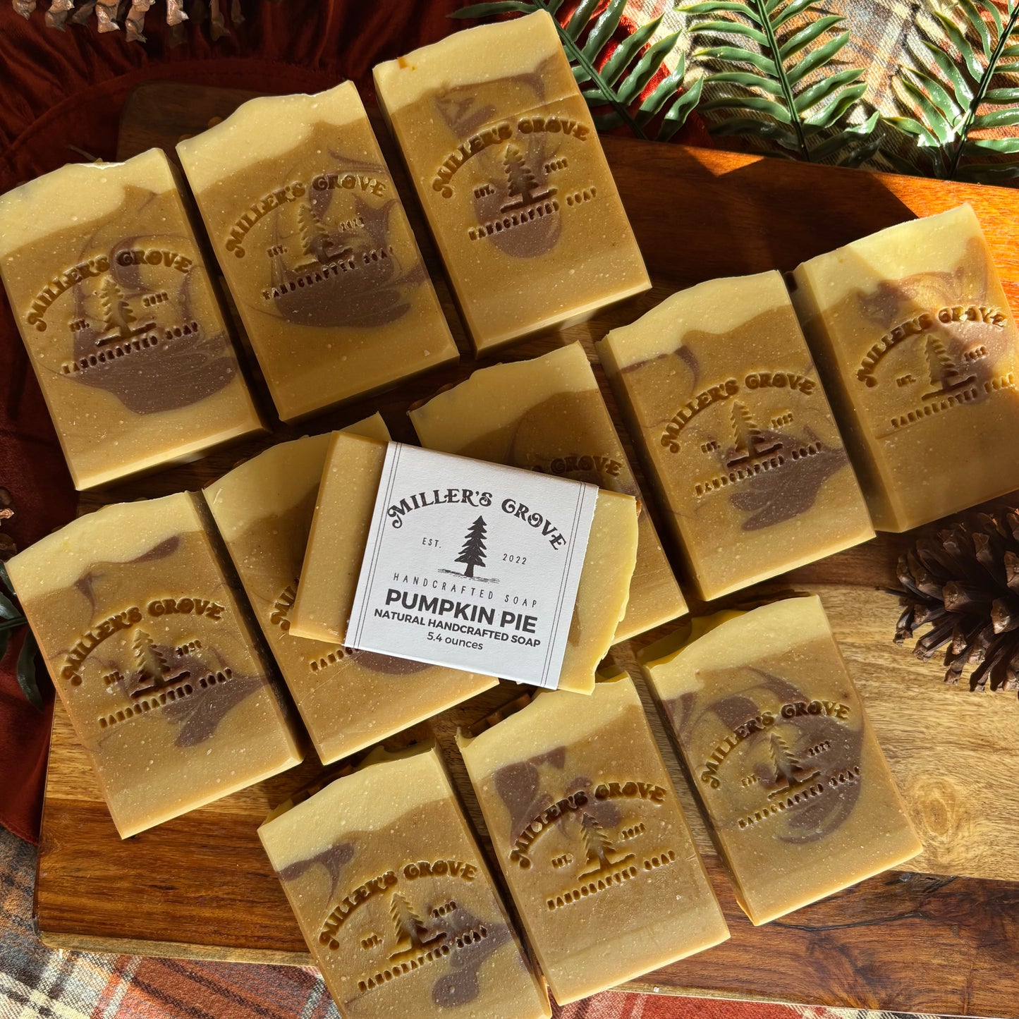 Nine bars of Miller's Grove handmade soap, shaped like pumpkin pie slices with a white paper wrapper around the center bar, arranged on a wooden board with pinecones and a plaid cloth.