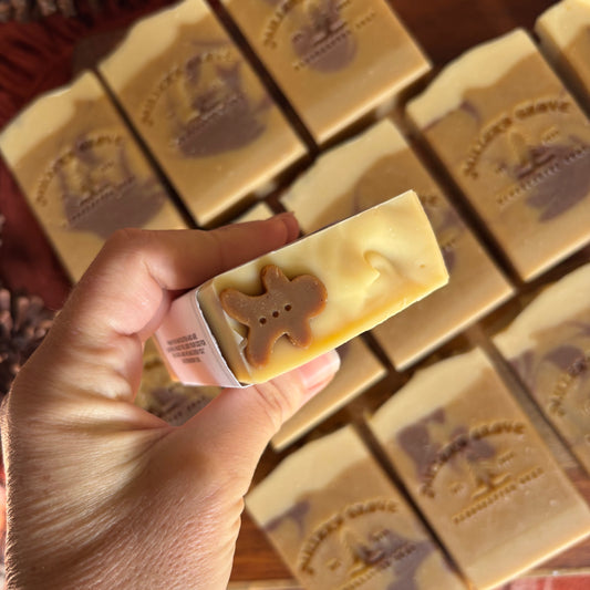 A hand holds a rectangular bar of soap in the foreground, with several matching soap bars arranged in the background. The held soap is pale cream with a brown gingerbread man shape molded into one end.